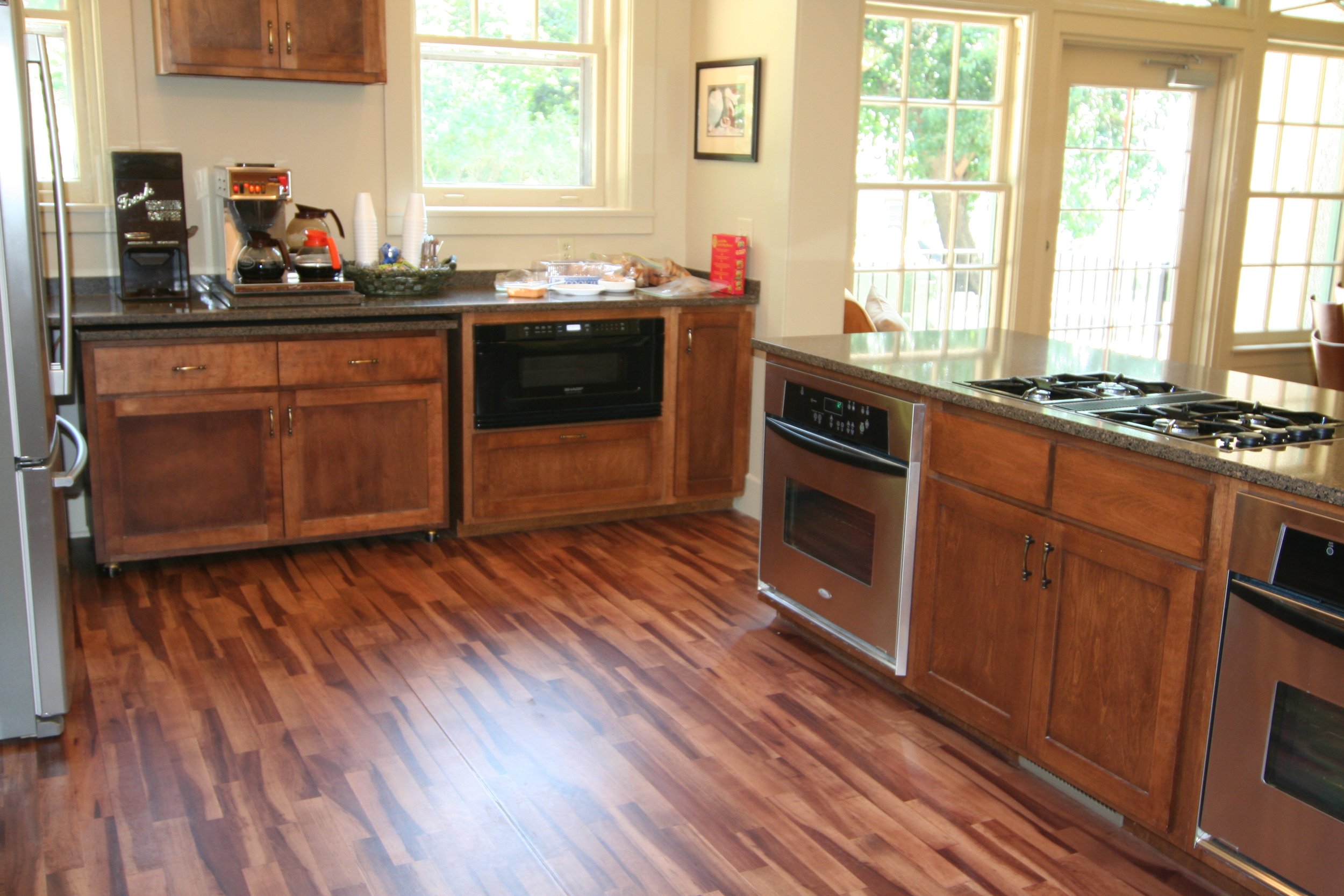 A kitchen with wooden cabinets, black countertops, and stainless steel appliances, including a stove, oven, coffee maker, and refrigerator. There are windows with a view of greenery outside and a wooden floor.