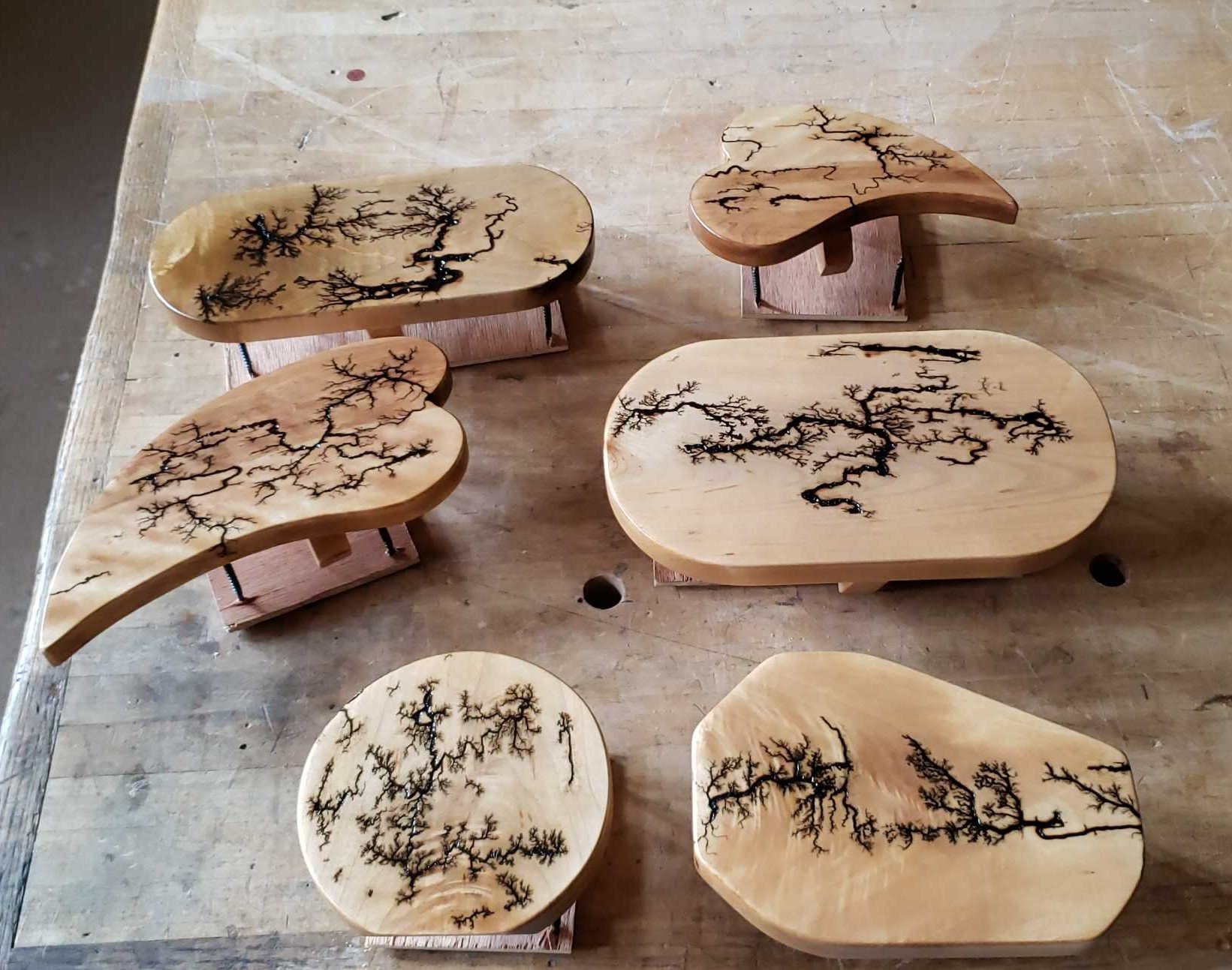 A collection of six wooden tables with black tree branch designs burned or painted onto their surfaces, arranged on a wooden workbench.