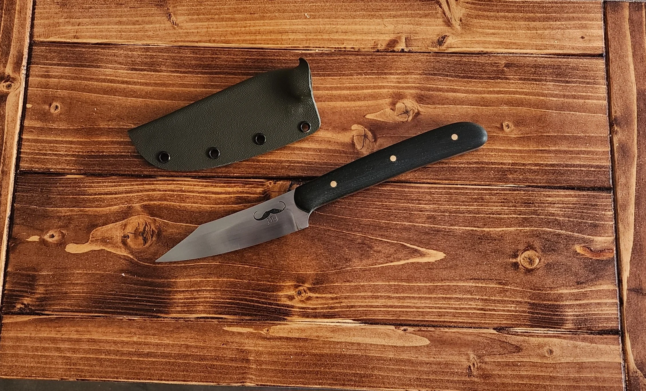 A small tactical knife with a black handle and a stainless steel blade, accompanied by a dark green sheath, resting on a wooden surface.