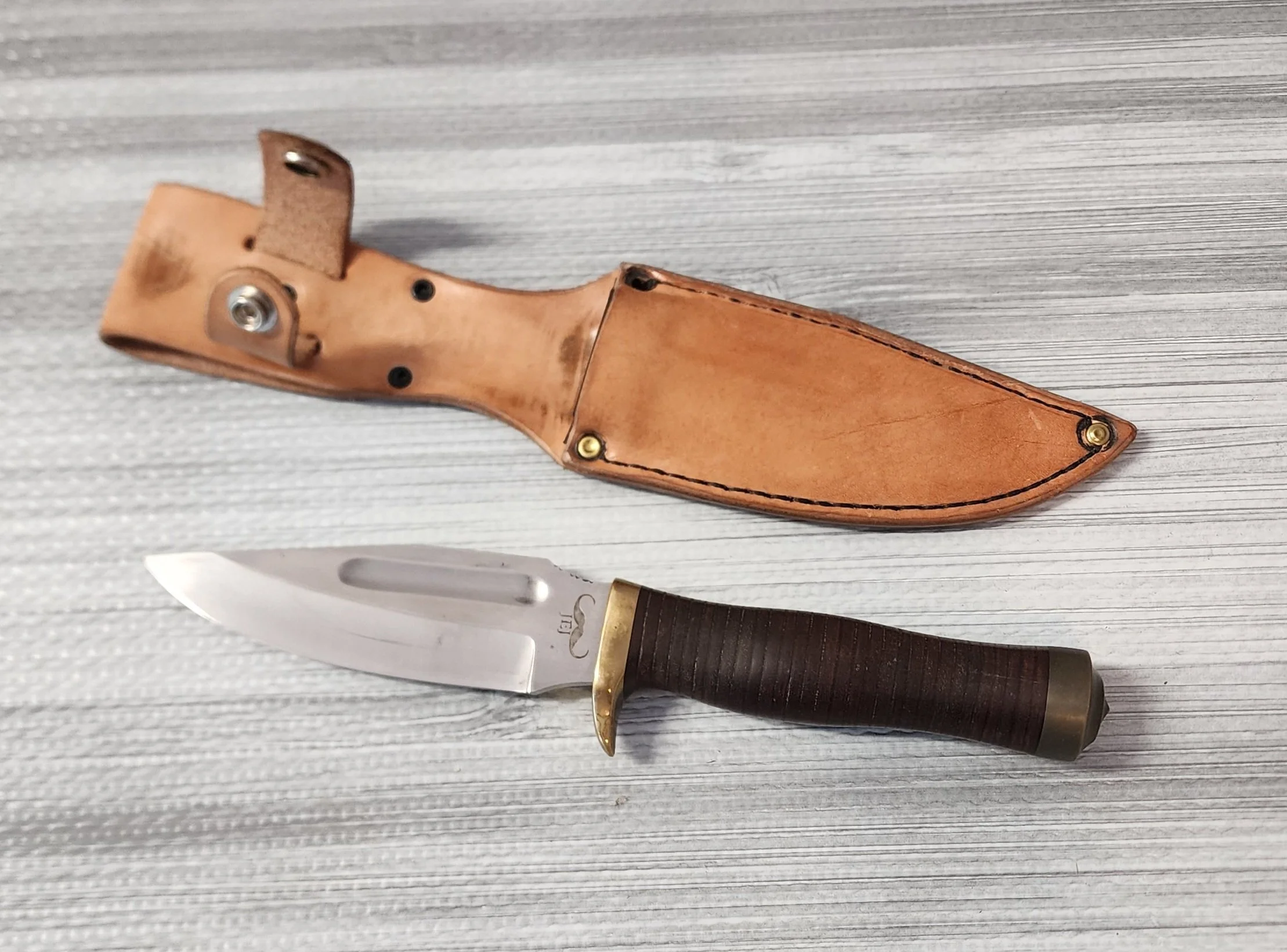 A hunting knife with a dark wooden handle and brass guard, along with a leather sheath, both placed on a light gray textured surface.