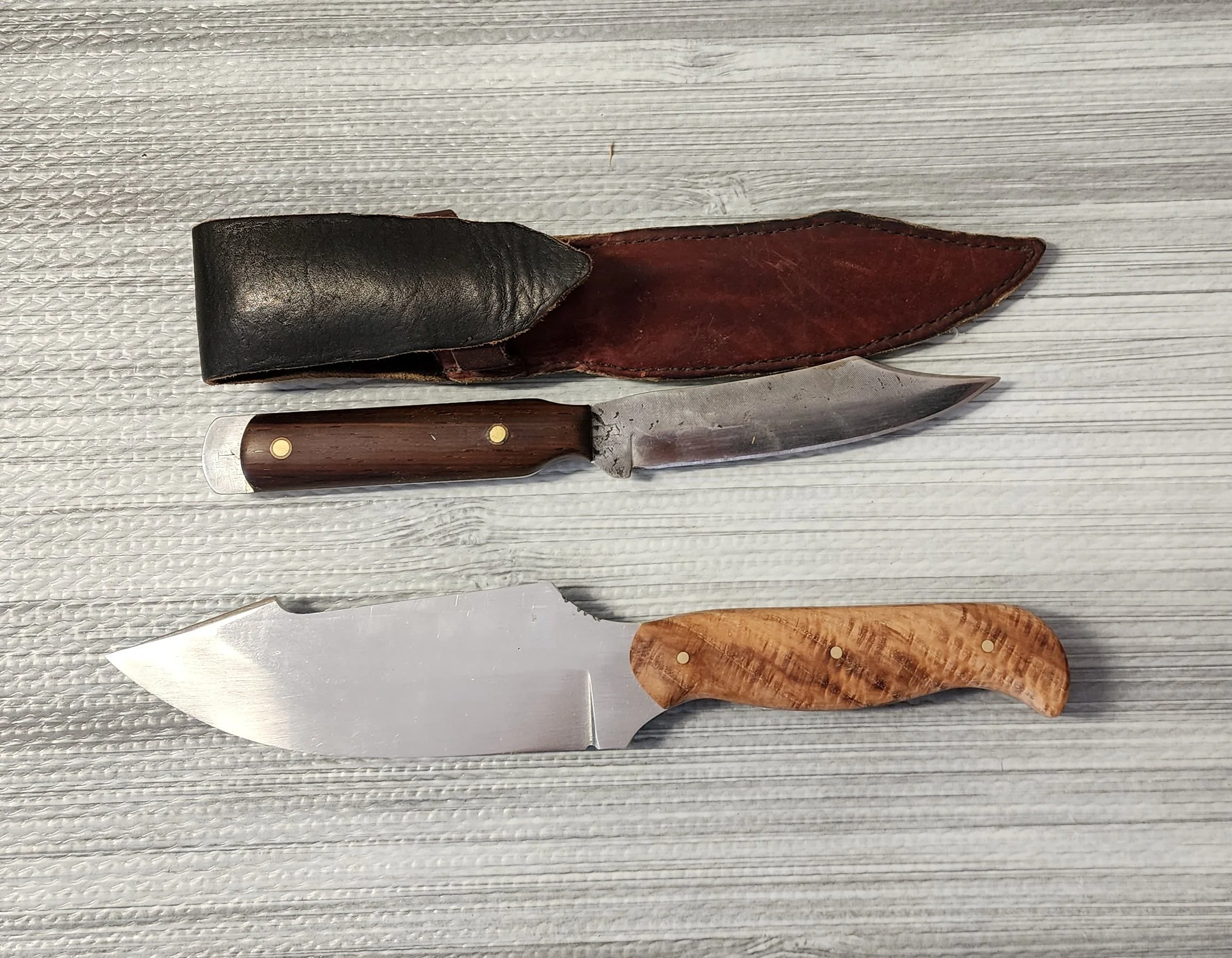 Two knives with wooden handles, one with a black leather sheath and the other without, placed on a light gray wooden surface.