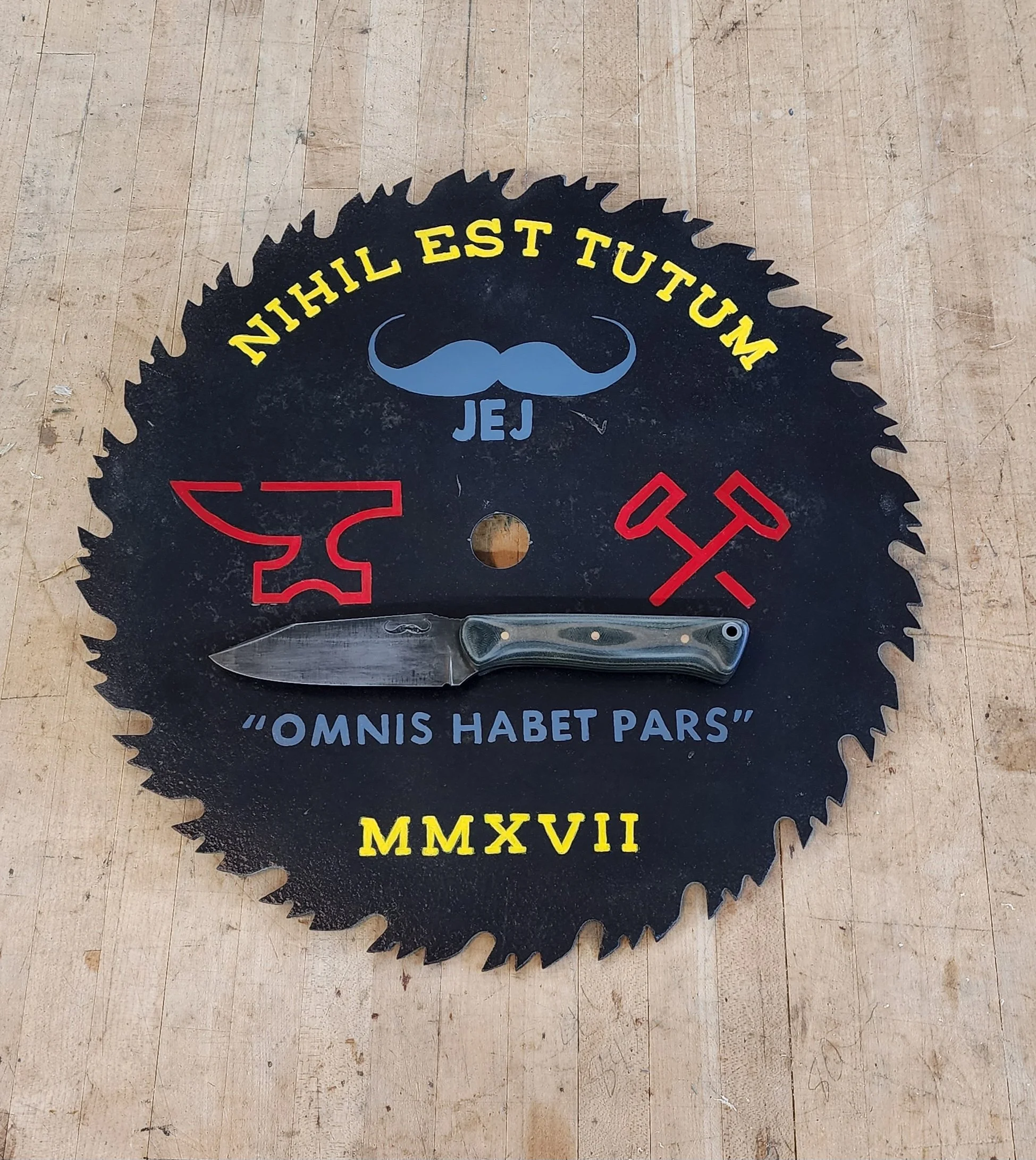 A circular saw blade decorative piece with various symbols, text, and a knife placed on top. The saw blade has a quote in Latin, 'OMNIS HABET PARS,' and Roman numerals 'MMXVII,' along with Latin and other phrases, mustached face illustration, and sym