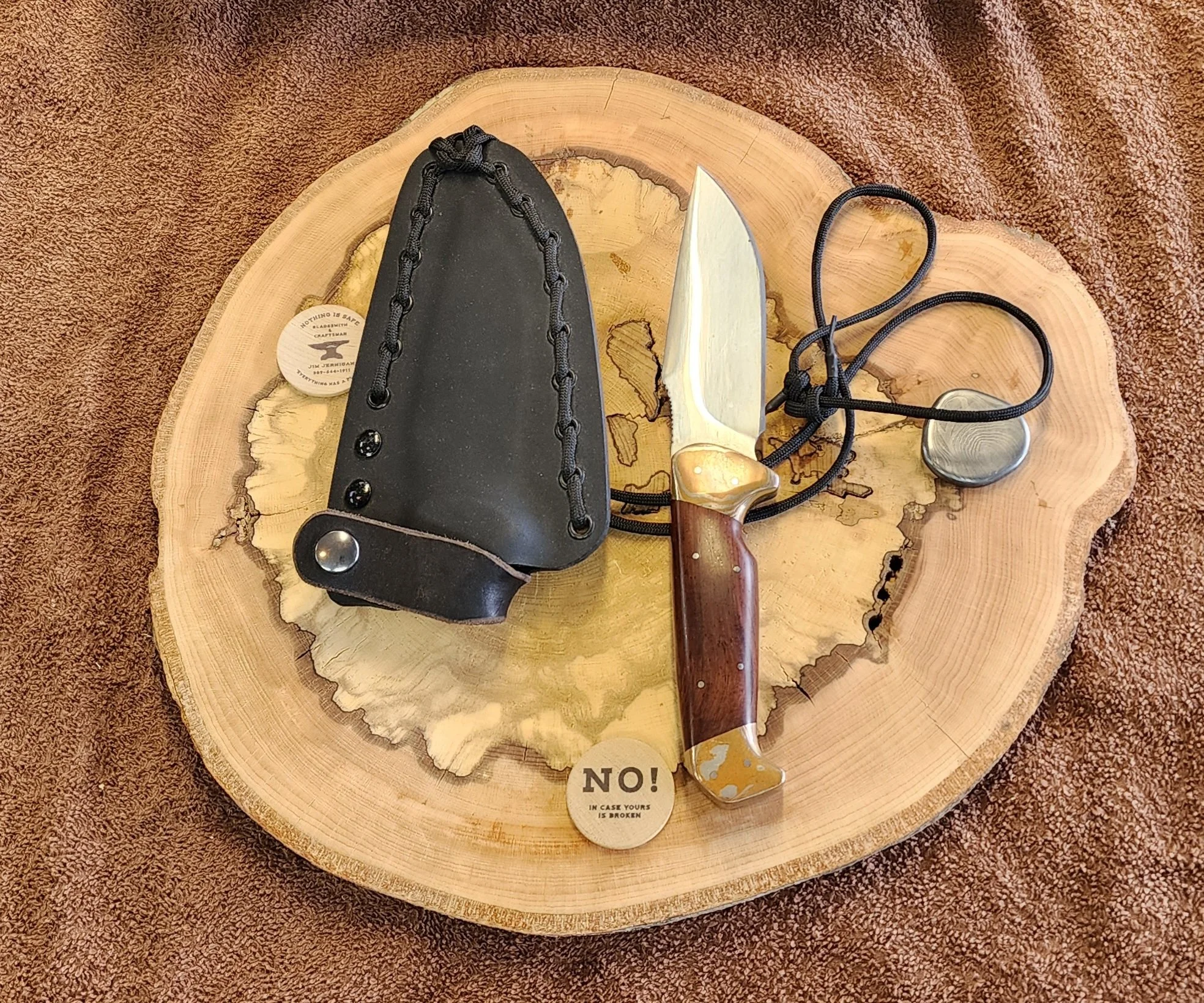 A fixed-blade knife with a wooden handle, a leather sheath, a stone, and a small token that says 'NO! IN CASE YOURS IS BROKEN' are arranged on a round wooden tree slice on a brown textured fabric surface.