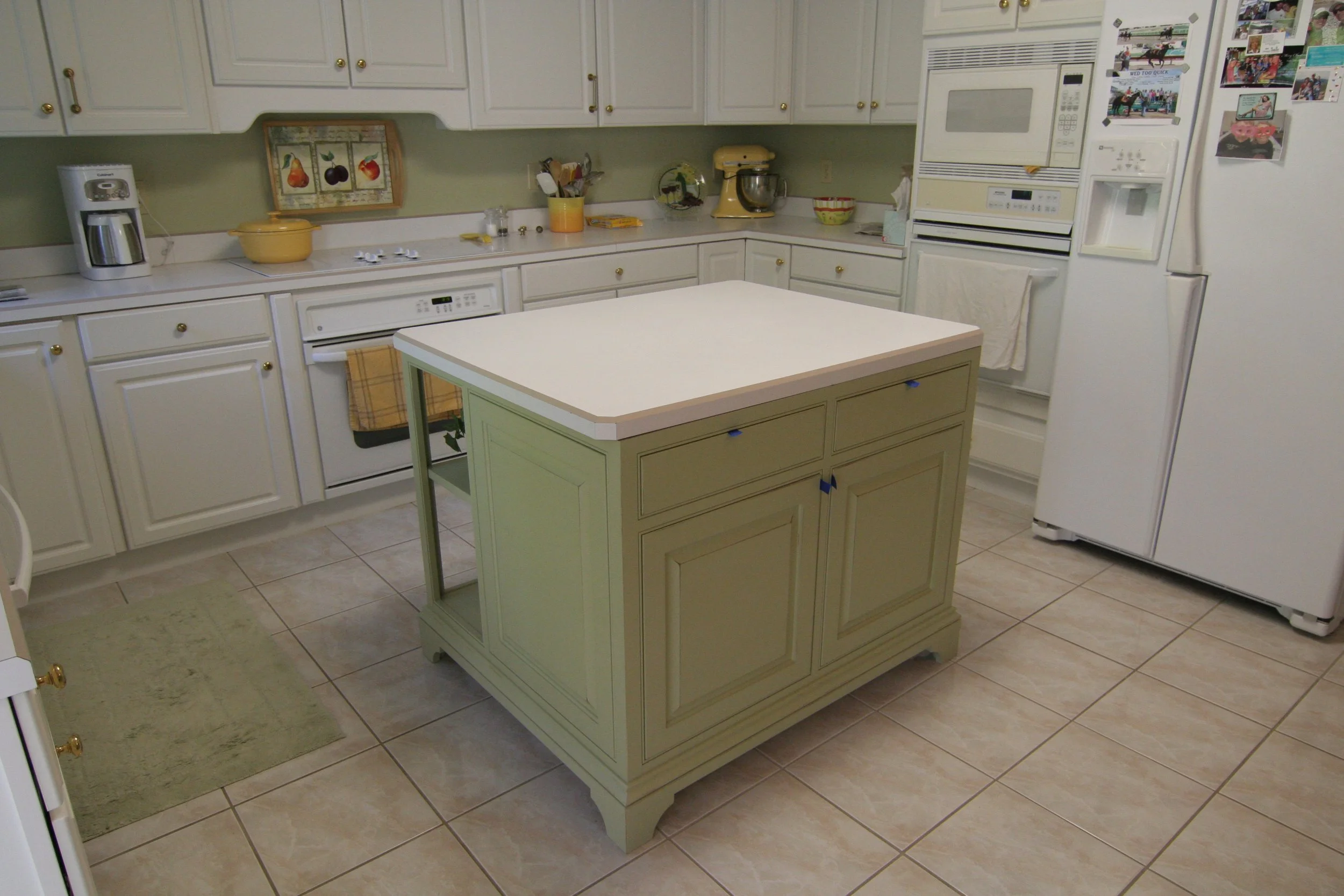 A kitchen with white cabinets, a white refrigerator with photos, a kitchen island in sage green, and various small appliances and decorations.