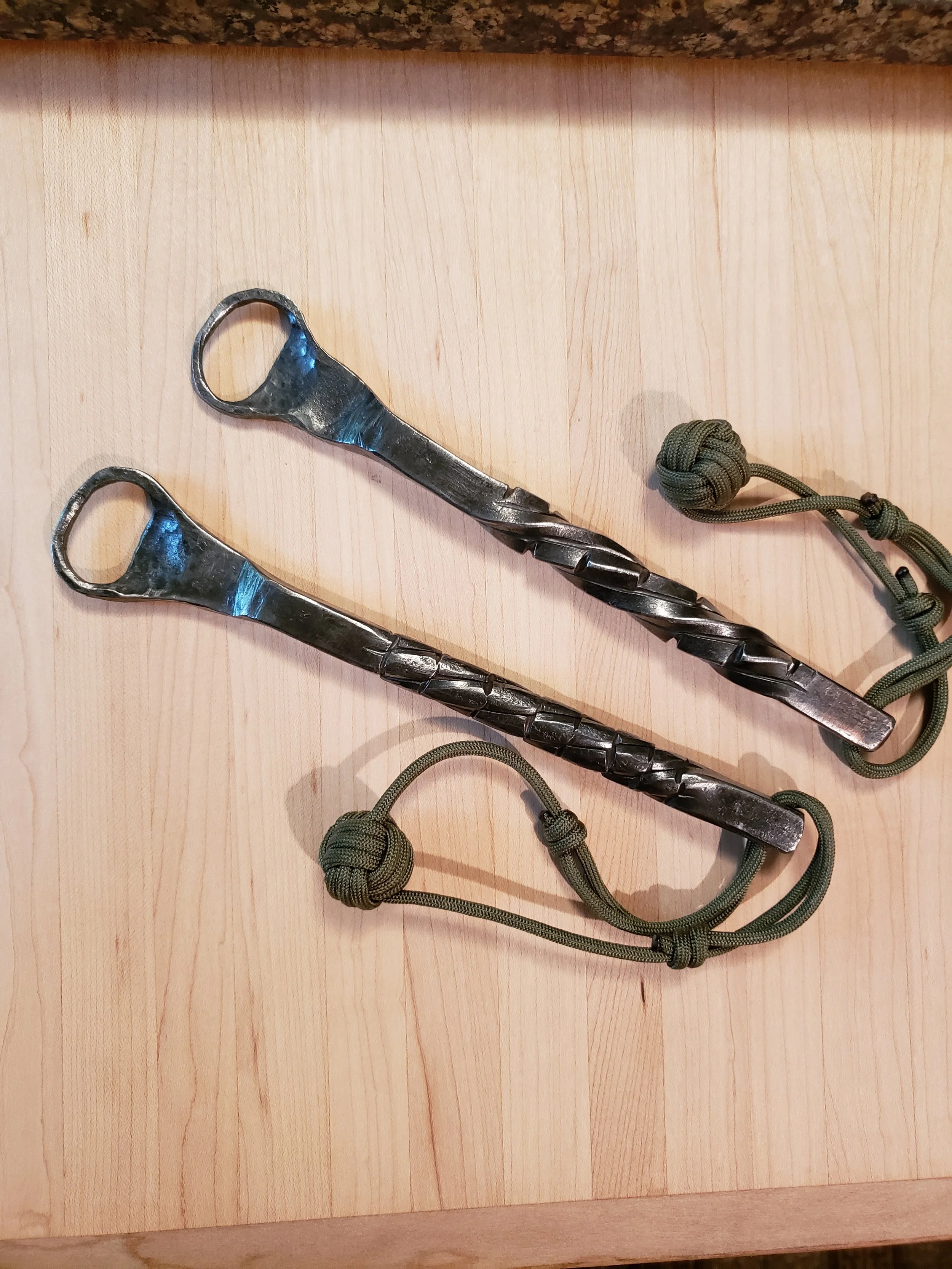 Two metal fire tools with a twisted design and attached green paracord lanyards resting on a light wood surface.