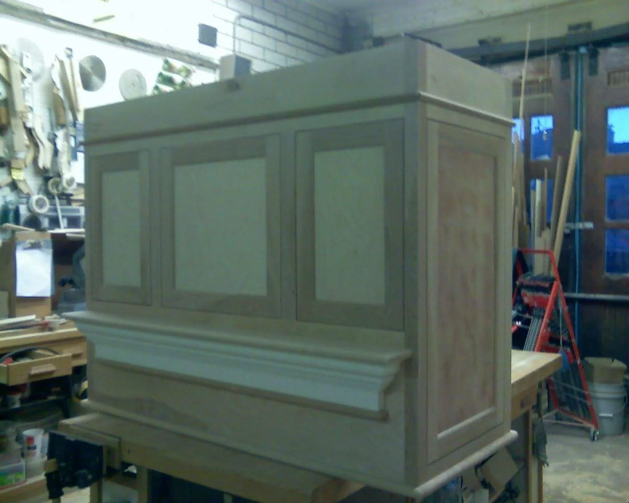 Unfinished wooden cabinet in a woodworking workshop with tools and supplies visible in the background.