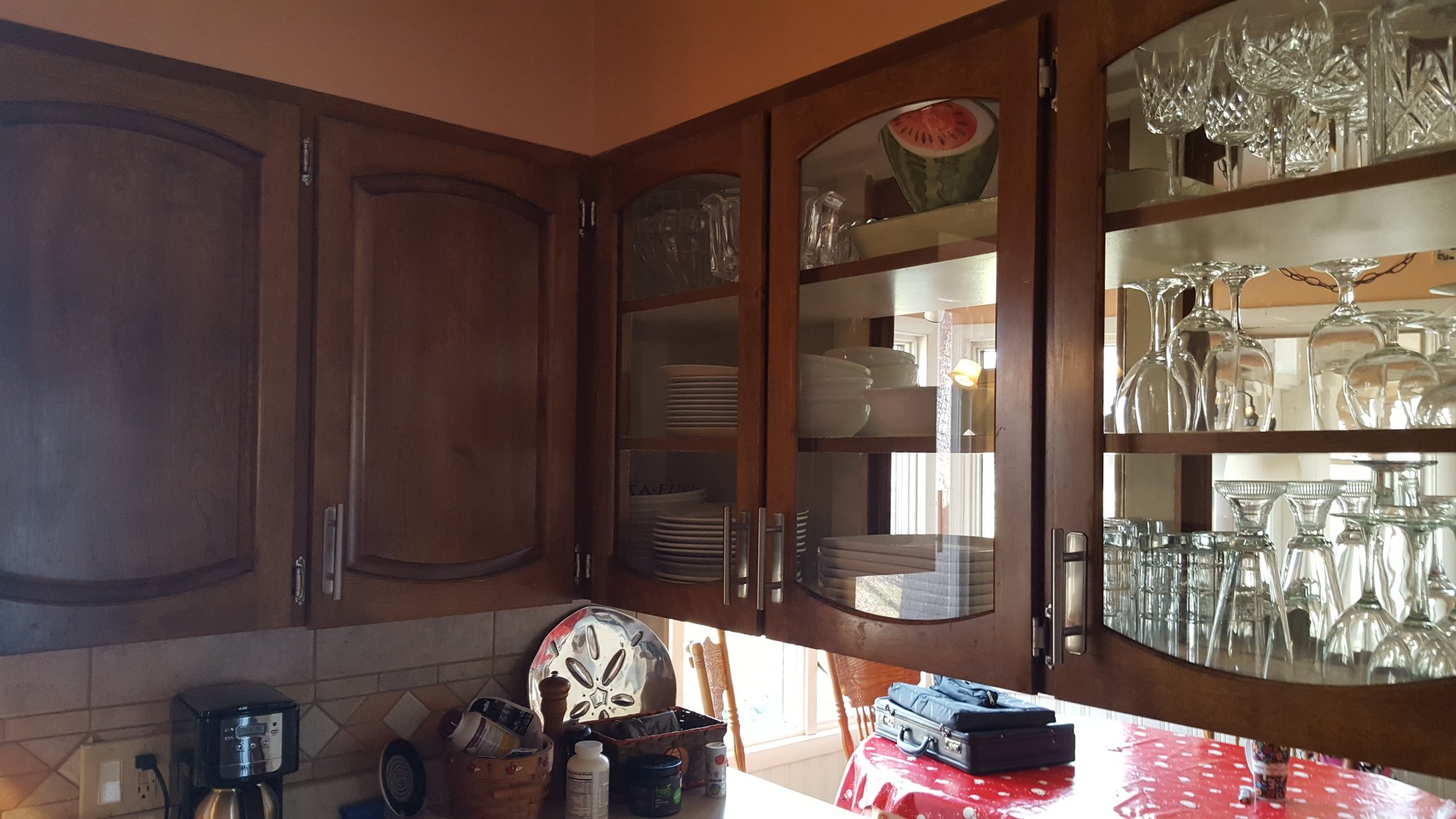 Kitchen cabinet with glassware and dishes, a coffee maker, a table with a red polka-dot tablecloth, and various items on the counter.