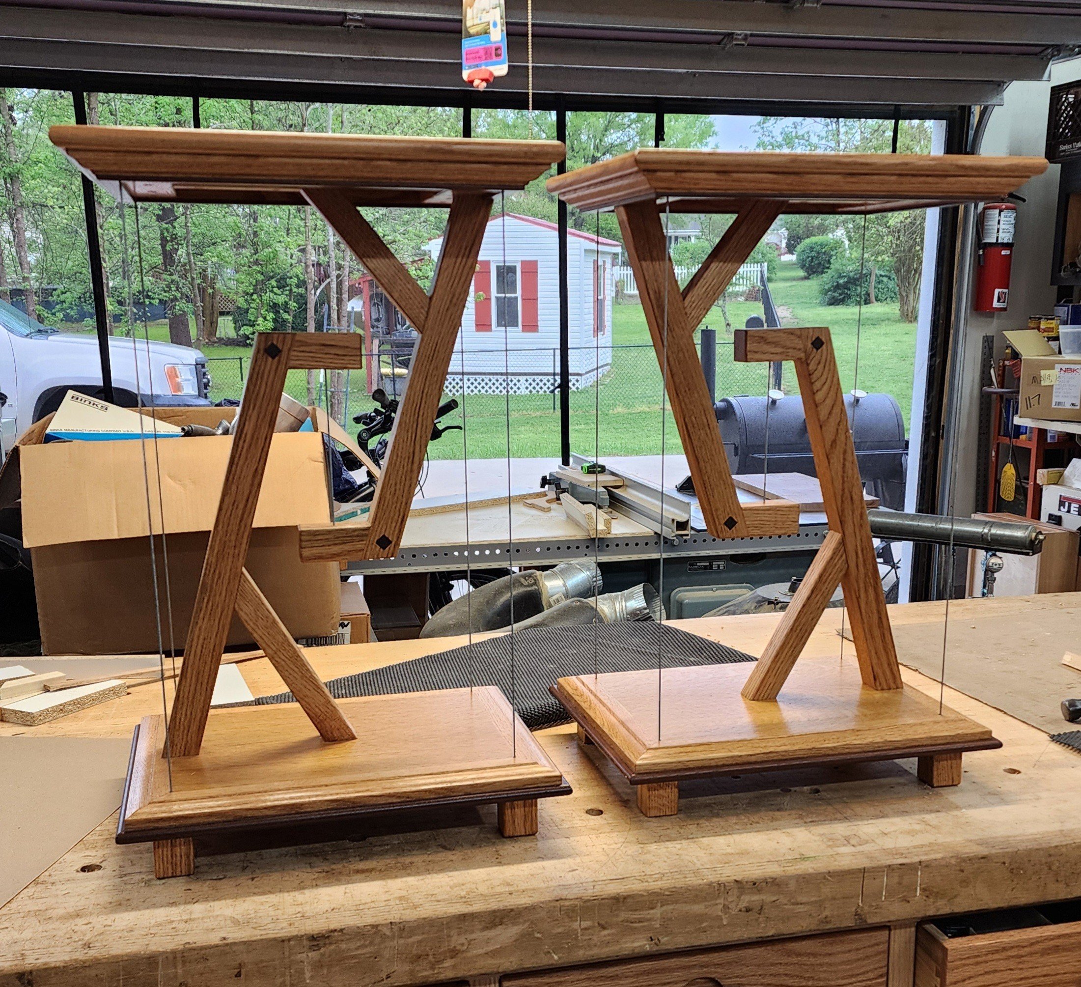 Two wooden stands with glass panels in a woodworking shop, with a garage door and yard visible in the background.