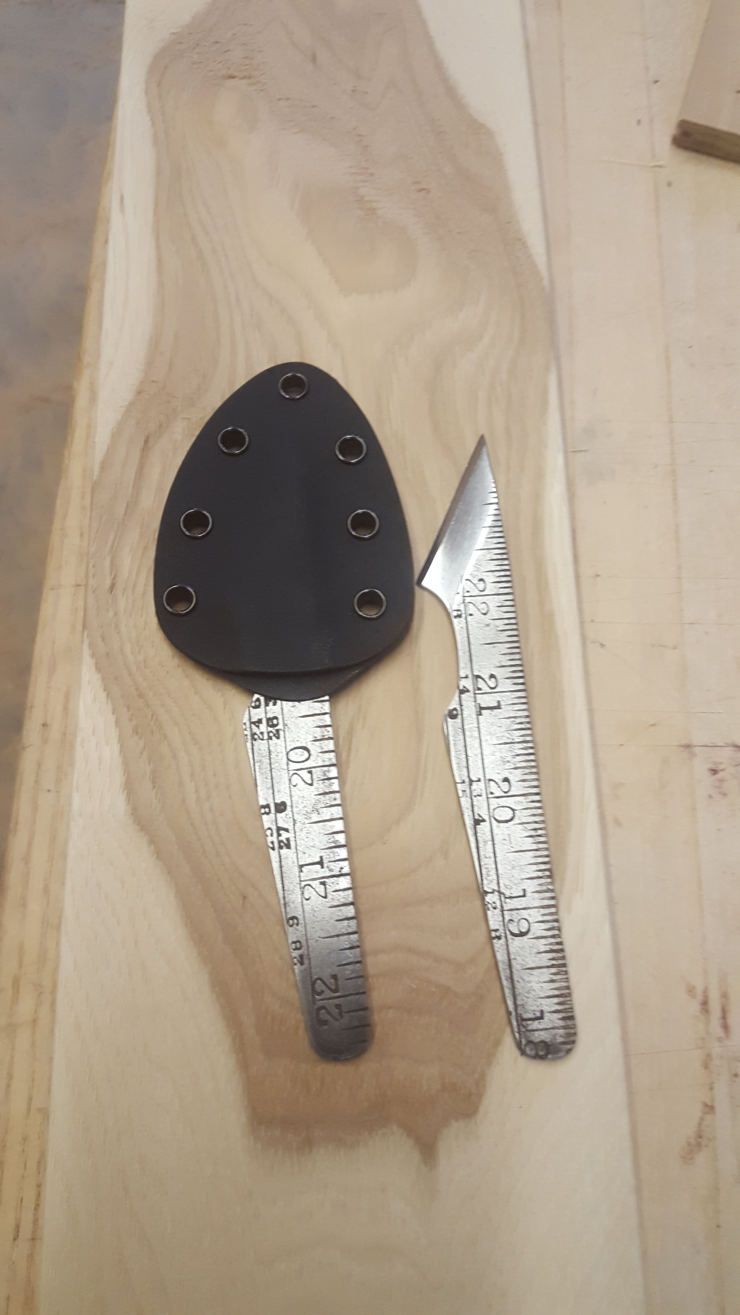 A black guitar pick with six small metal rings on it, placed on a piece of wood, with two metal rulers next to it for scale.