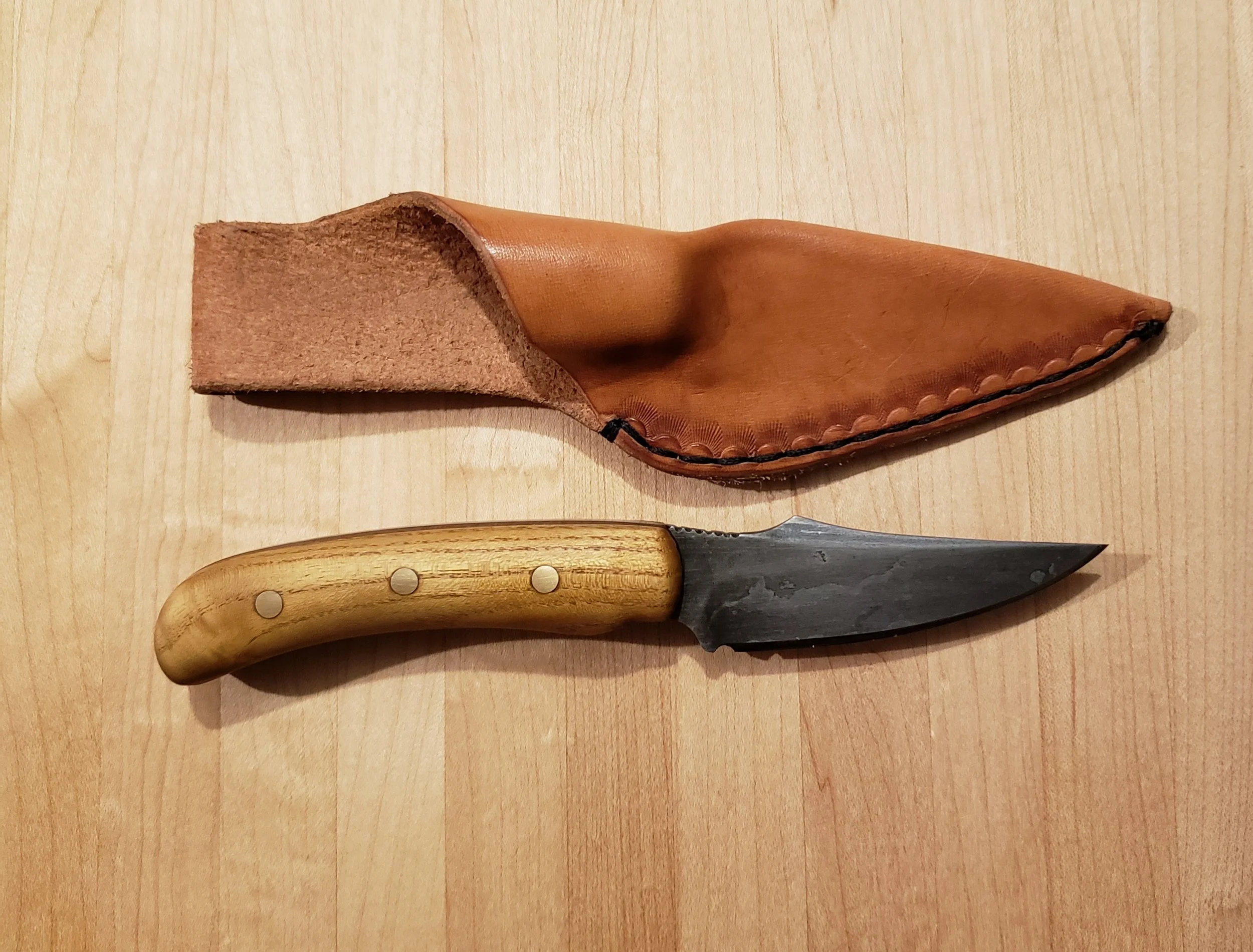 A small black fixed-blade knife with a wooden handle resting on a light wooden surface. The knife has a slightly curved blade. There is a brown leather sheath, shaped to fit the knife, with a stitched edge, positioned above the knife.