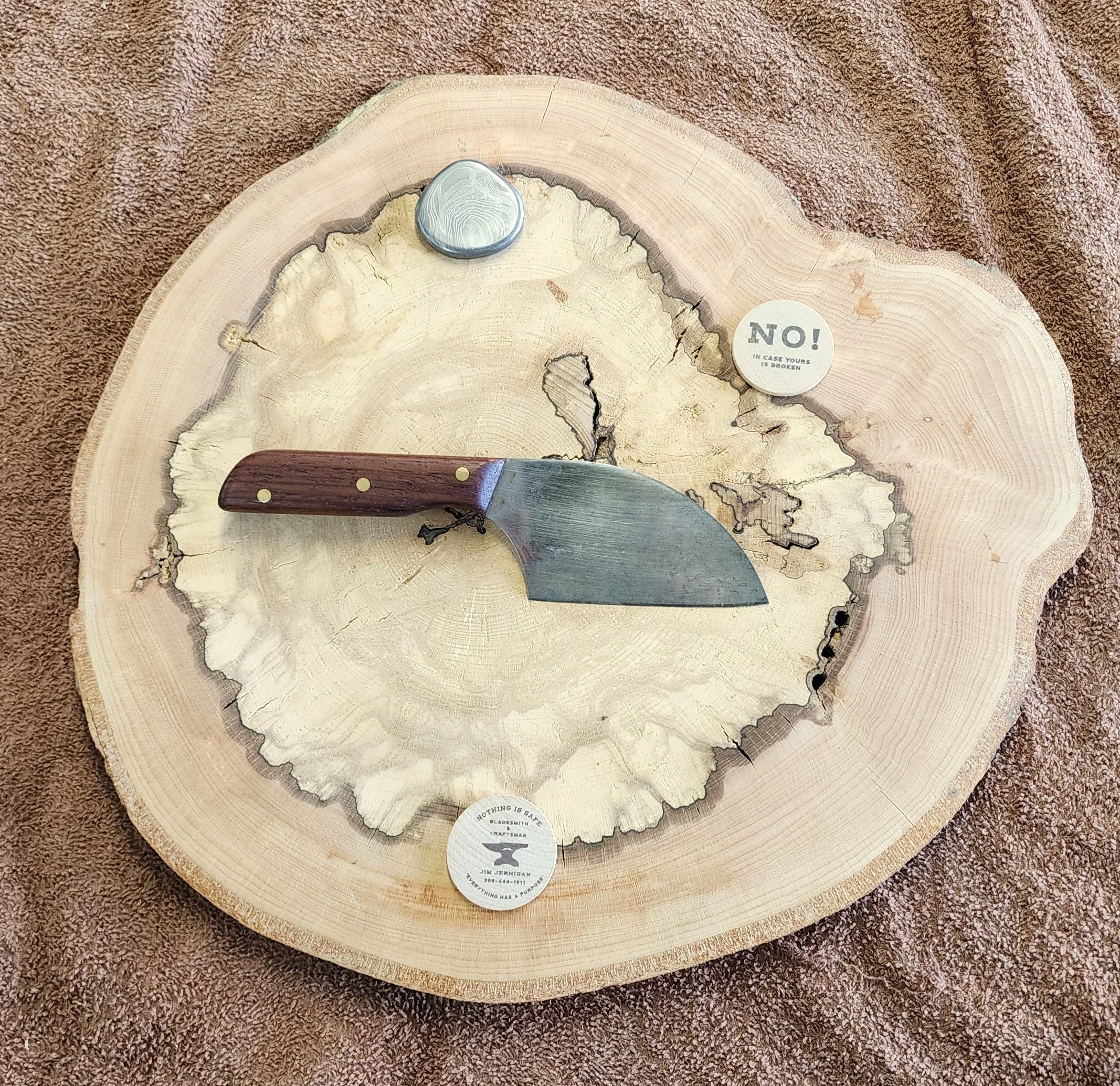A wood slice with a small cleaver on top, and three circular pins or tokens on the surface, one of which says 'NO! IN CASE YOURS IS BROKEN' and another says 'NOTHING IS SAFE' with additional text.