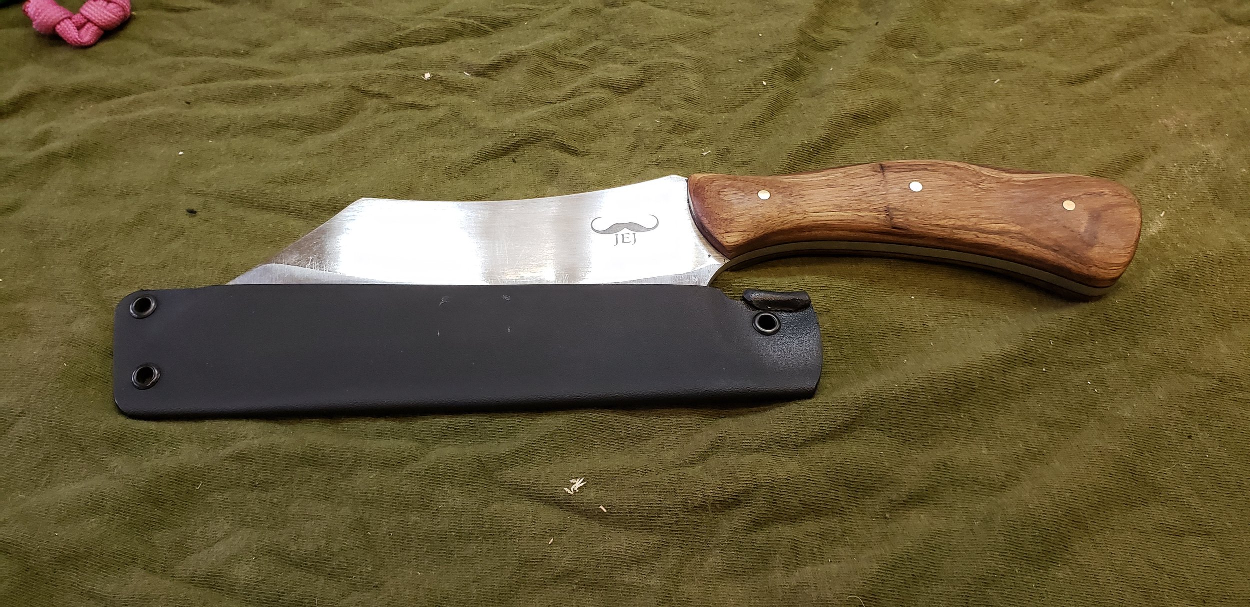 A knife with a wooden handle and a decorated blade, partially in a black sheath, lying on an olive green fabric background.