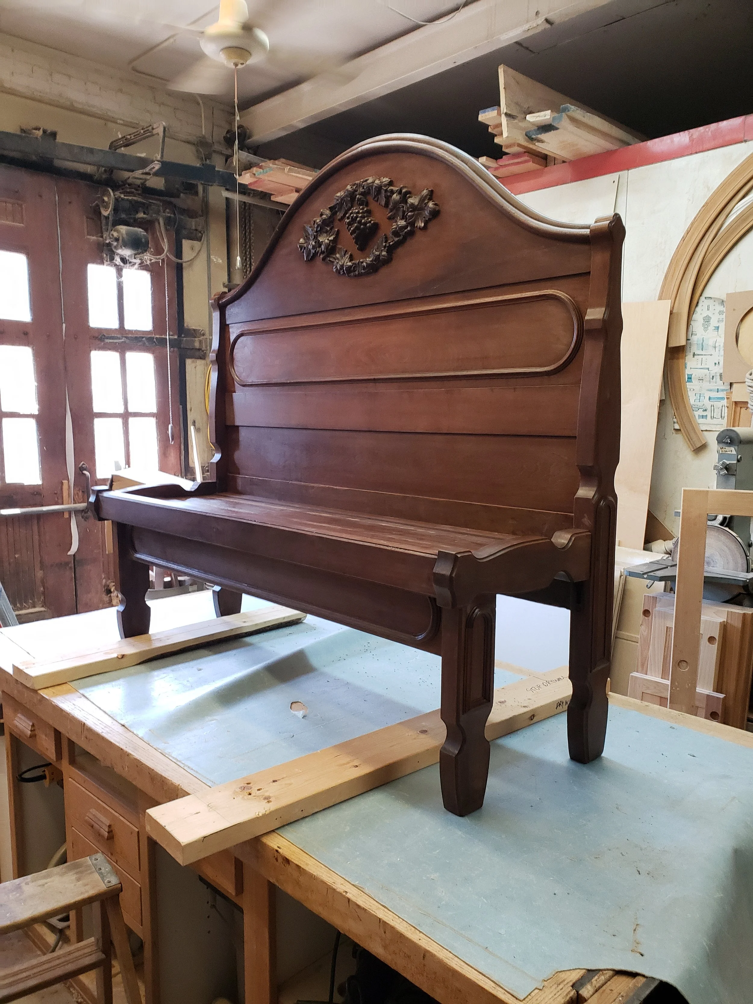 A wooden headboard with carved floral details, resting on a worktable in a woodworking shop.