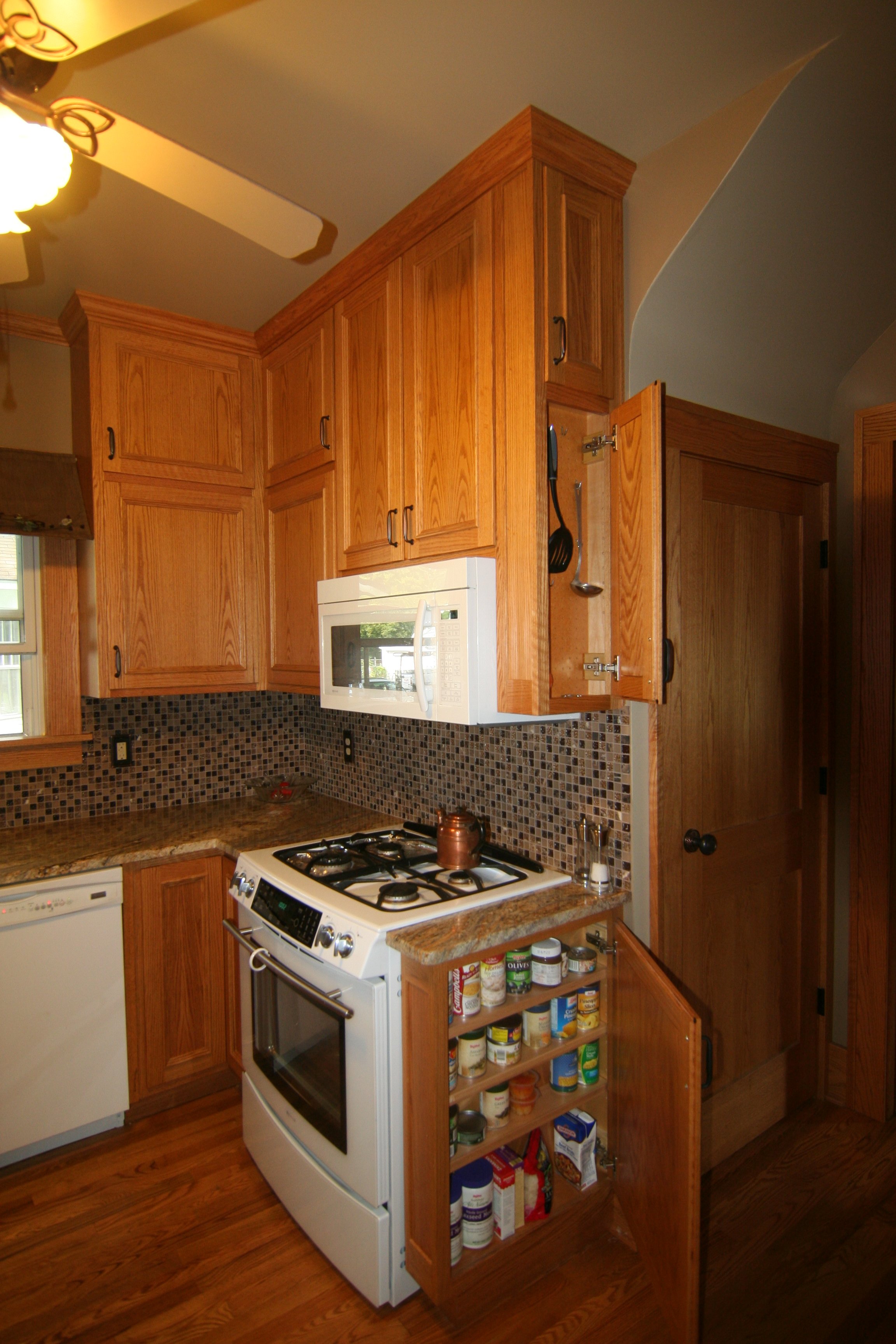 Kitchen with wooden cabinets, microwave, stove, spice rack, and pantry with open door showing canned goods.