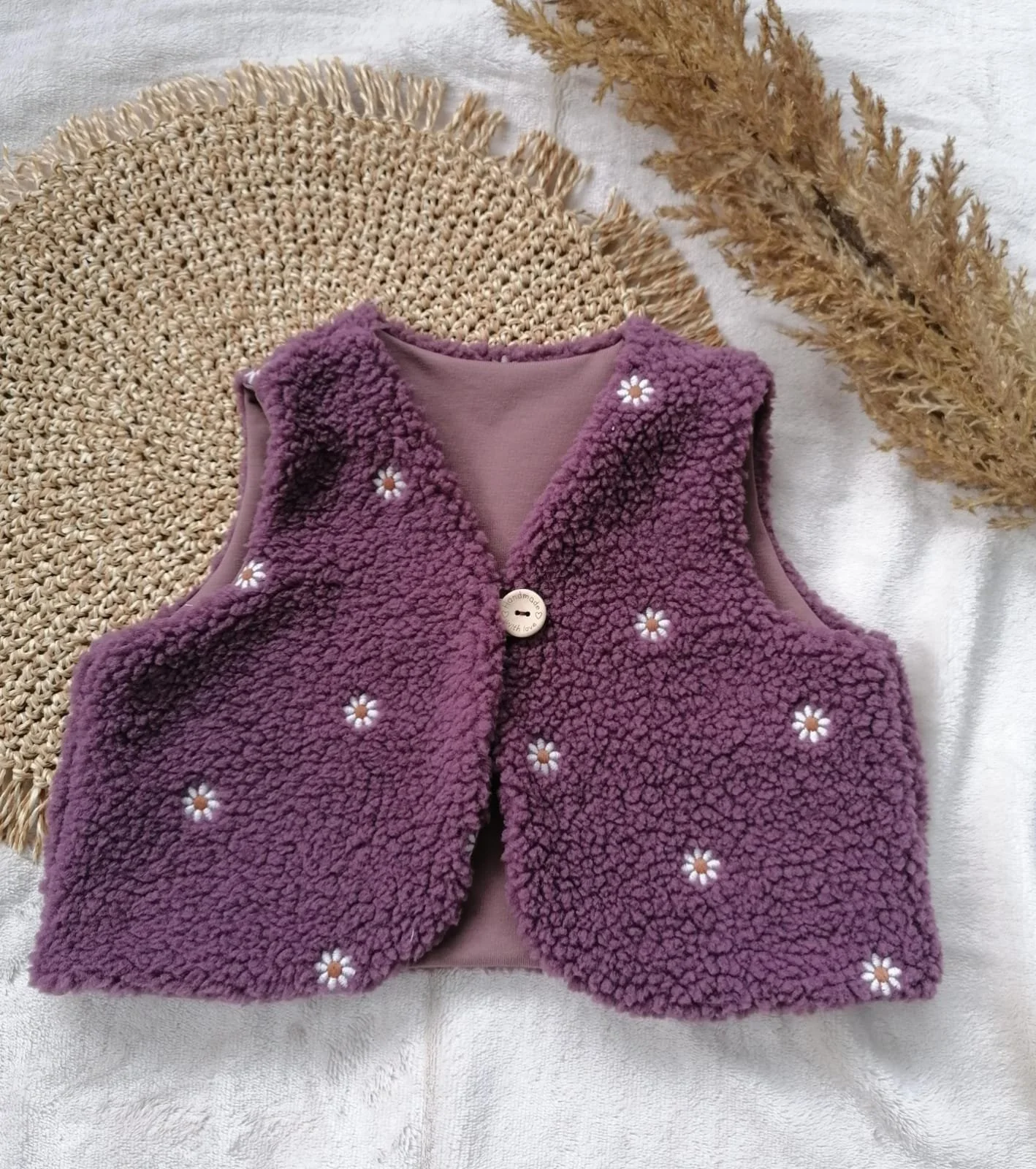 Lila kuschelige Weste mit kleinen weißen Blumen mit gelben Zentren, auf einer weißen Decke, daneben getrocknetes Gras.