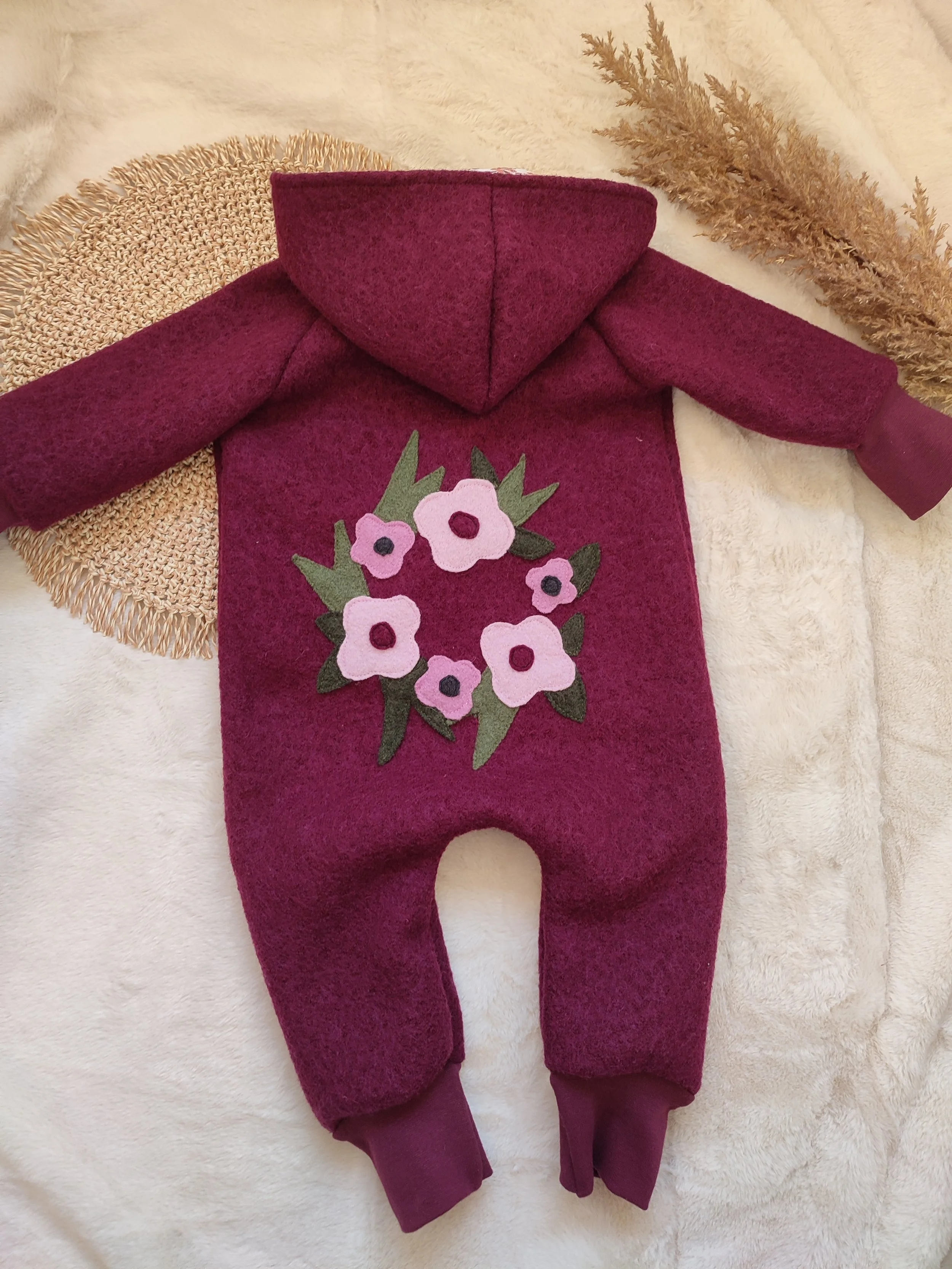Ein rot-violemer Babystrampler mit Kapuze und einer Blumendekoration auf der Rückseite. Das Kleid liegt auf einem cremefarbenen, weichen Untergrund, umgeben von getrockneten Gräsern.