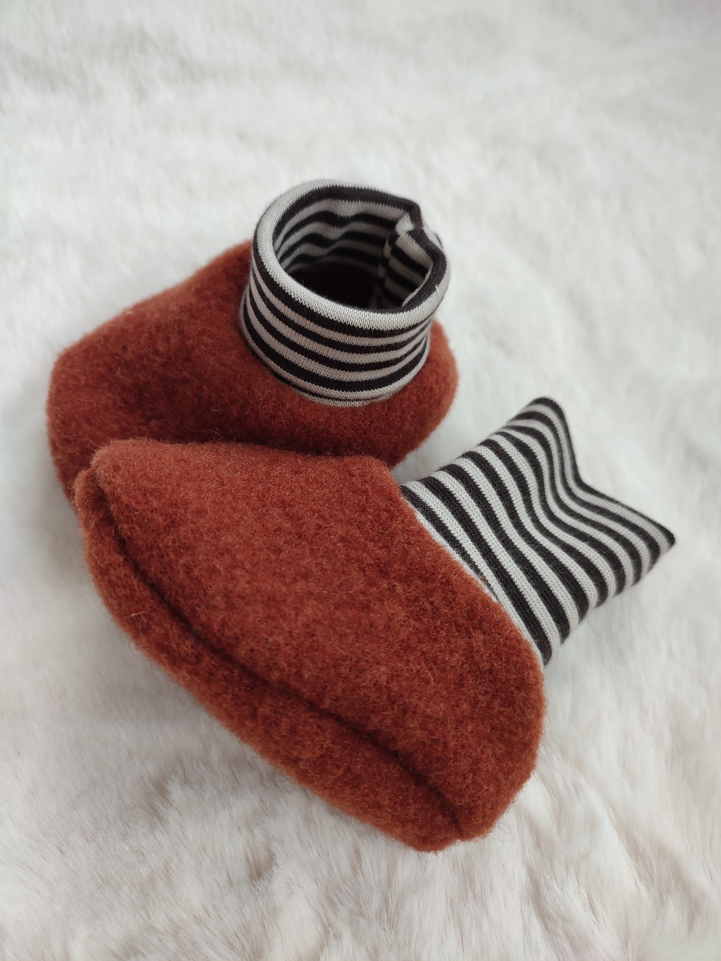 Ein gestrickter roter Babyschuh mit einem schwarzen und weißen gestreiften Kragen und Zunge, auf einem weißen, pelzigen Hintergrund.