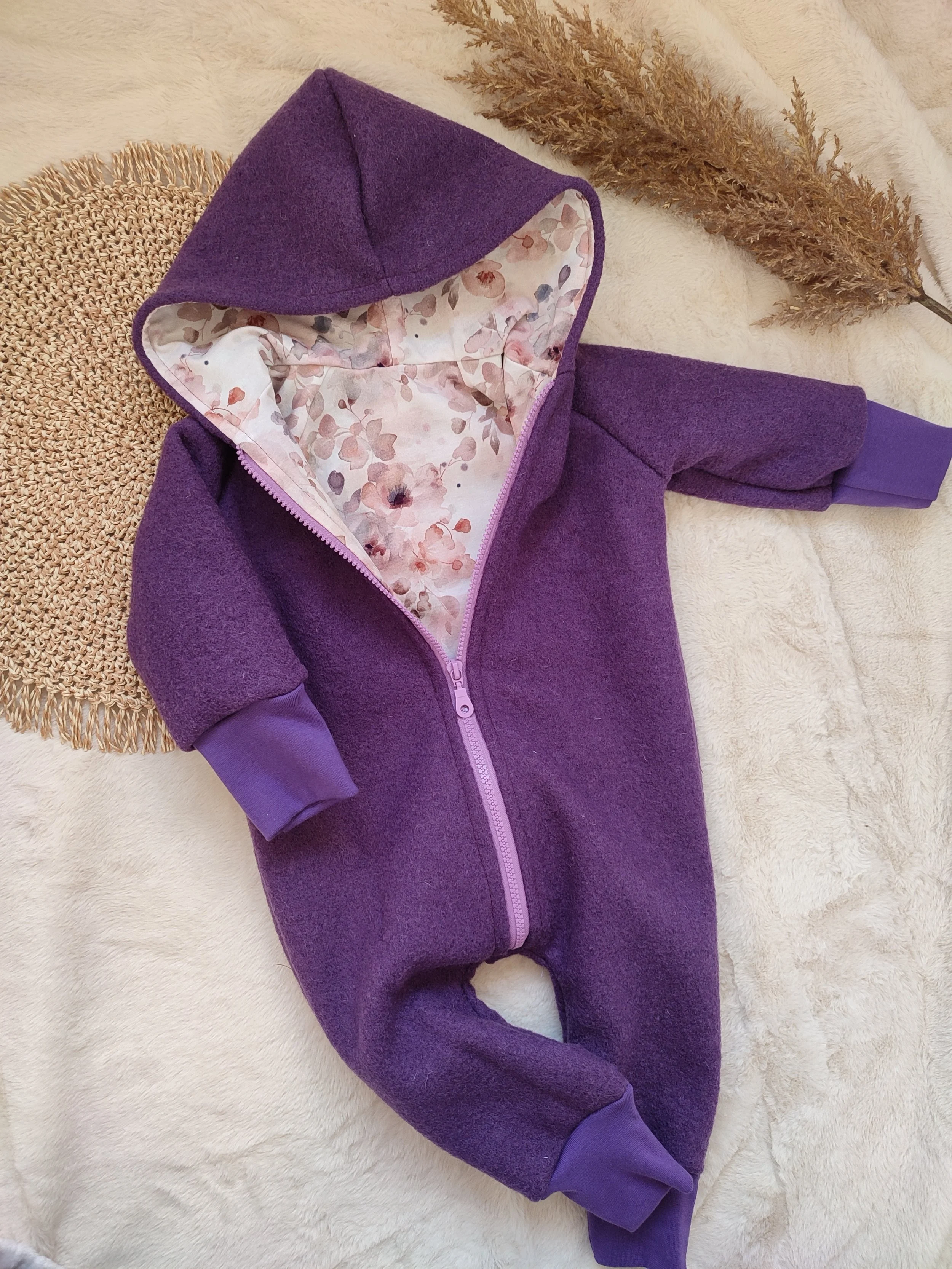 Ein lila Babystrampler mit Kapuze, offen mit floral bedrucktem Futter, auf einem weichen, hellen Stoff. Im Hintergrund befindet sich ein getrocknetes grasähnliches Dekor.