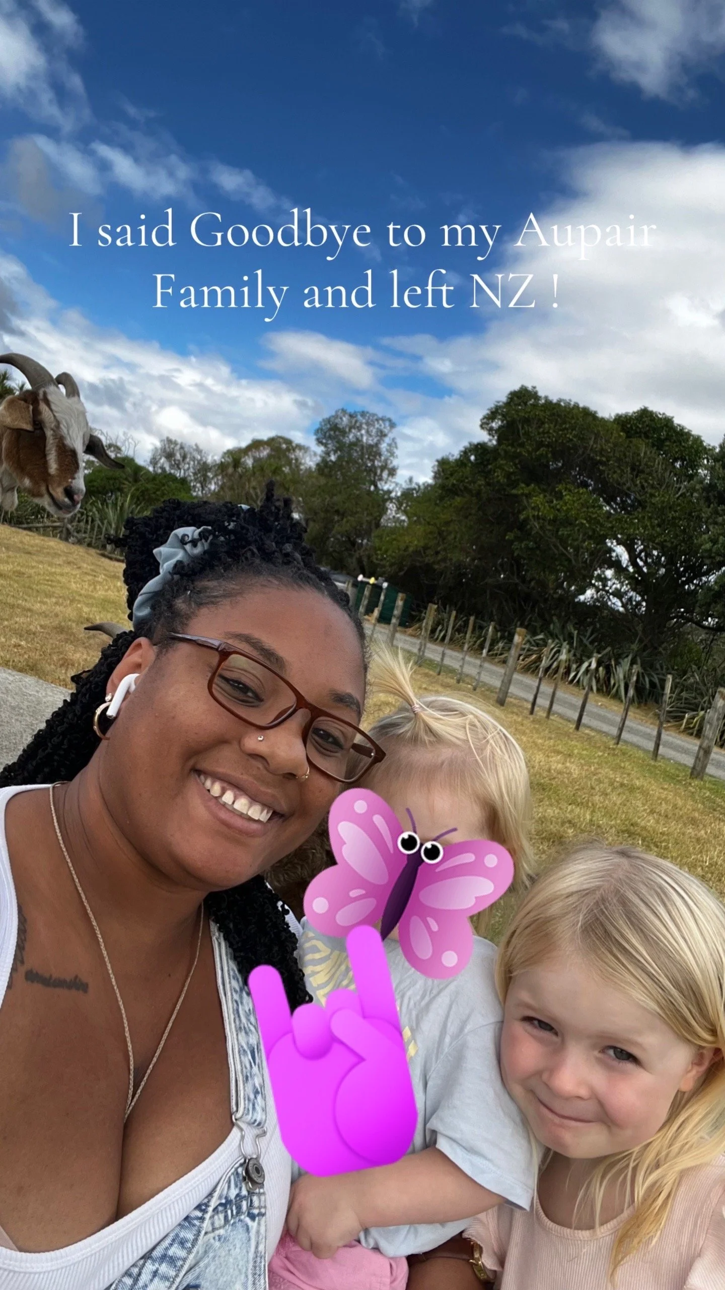 A woman with glasses and two children smiling outdoors with a goat in the background, trees, grass, and a blue sky with clouds.