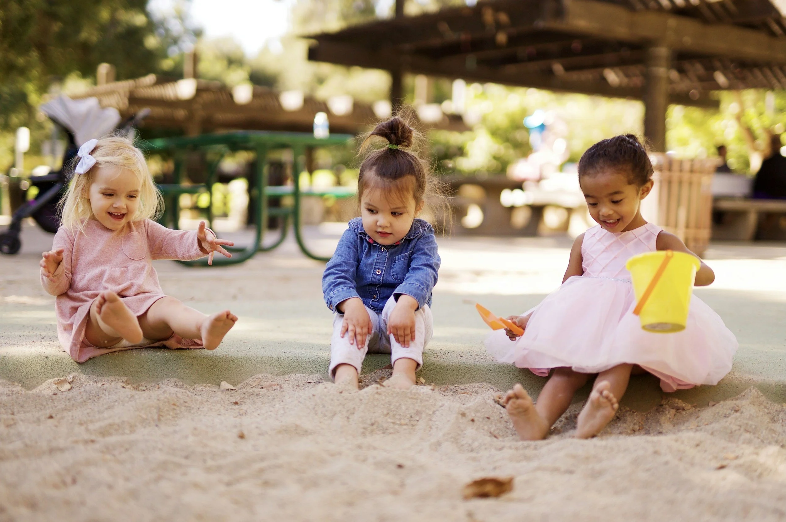 Three young girls playing in a sandbox at a park, surrounded by trees and park benches, enjoying outdoor play.