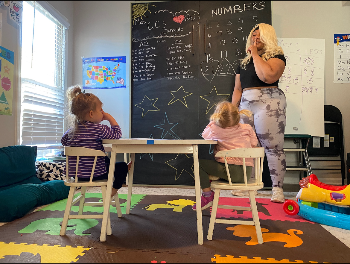 A young woman teaching children in a classroom with colorful foam mats, a chalkboard with numbers, schedule, and drawings, and a whiteboard with weather drawings. Two little girls sit at a small table, one in a striped purple shirt and the other in a pink jacket, listening to the teacher.