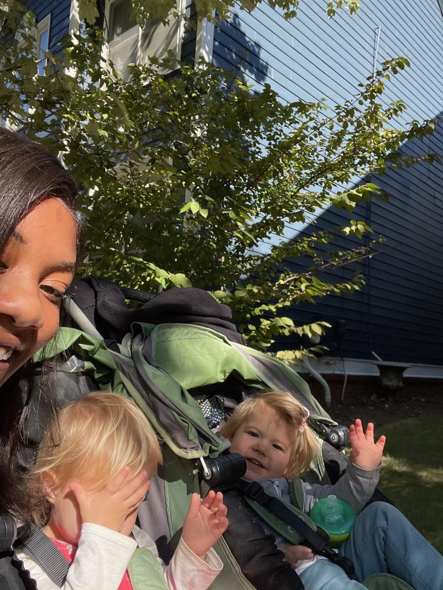 A woman and two children sitting outdoors on a sunny day, with a house and tree in the background. One child covers her face, and the other child is smiling and waving.