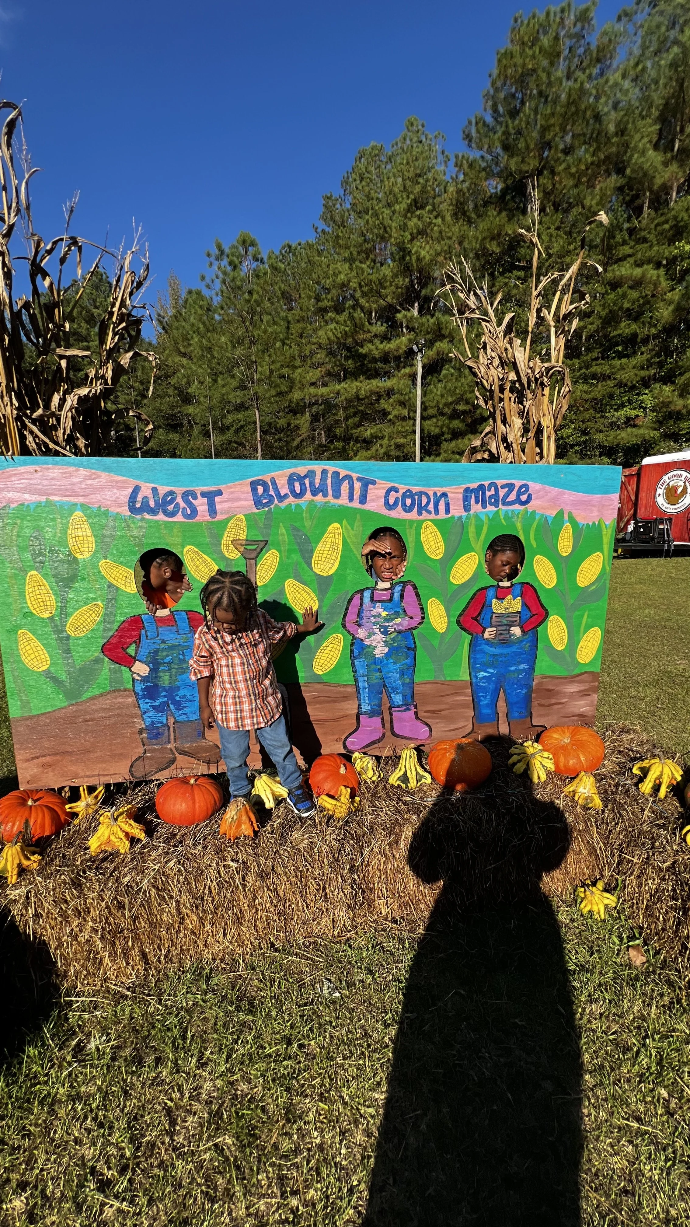 Children at a pumpkin patch stand with a colorful painted sign reading 'West Blount Corn Maze,' surrounded by pumpkins, gourds, and hay bales, with trees and a blue sky in the background.