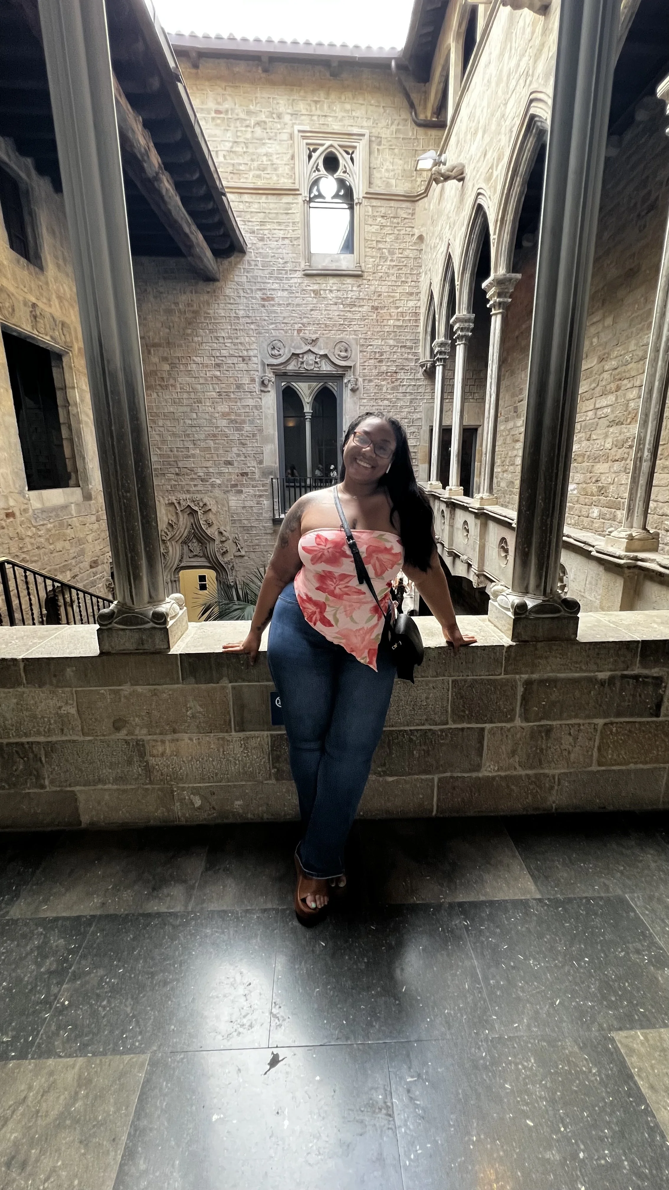 A woman smiling at the camera, standing in front of a stone railing in a historic building with gothic-style arch windows and stone columns.