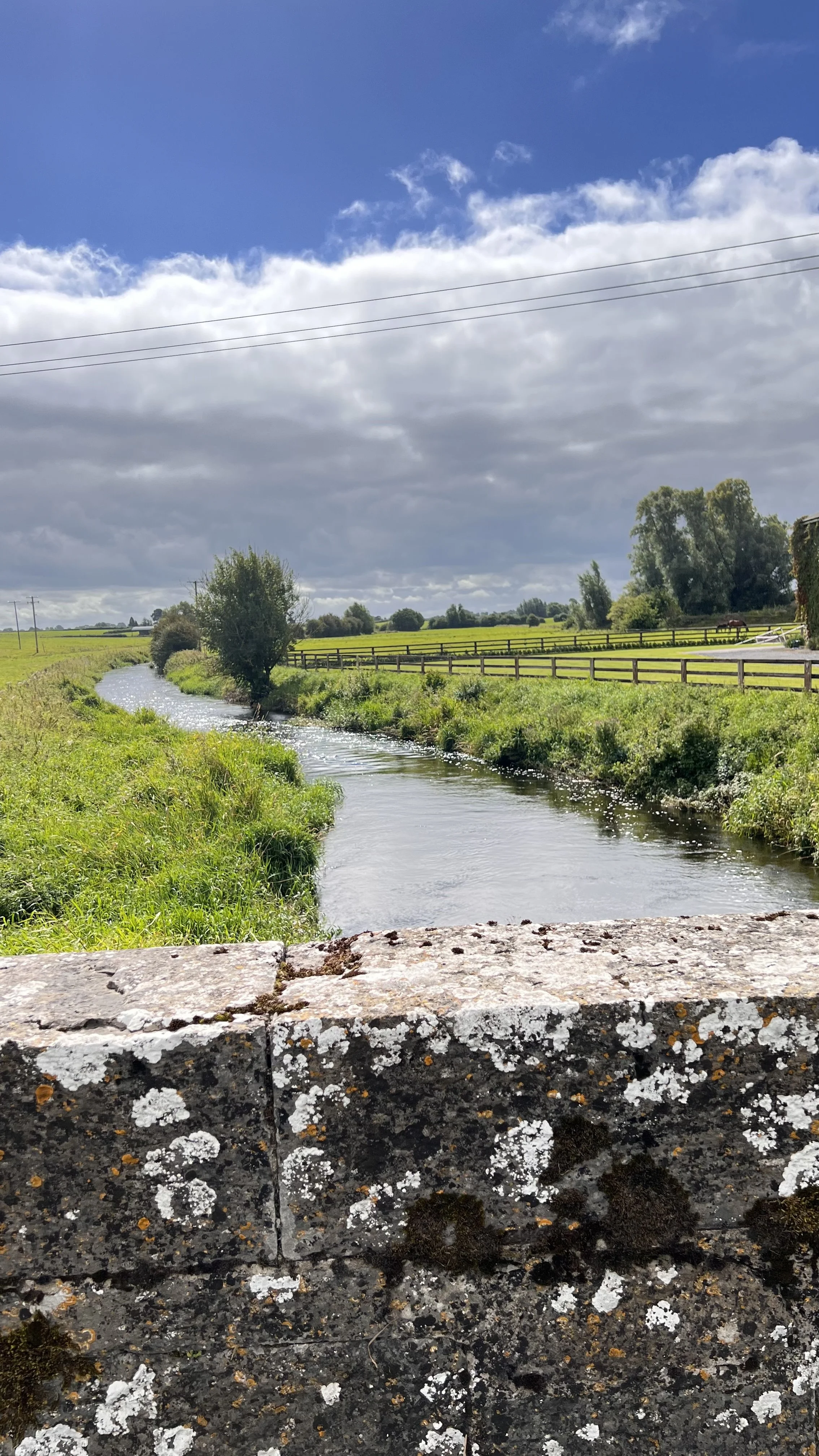 A scenic view of a small river flowing through lush green fields under a partly cloudy sky, with a stone bridge in the foreground and power lines overhead.