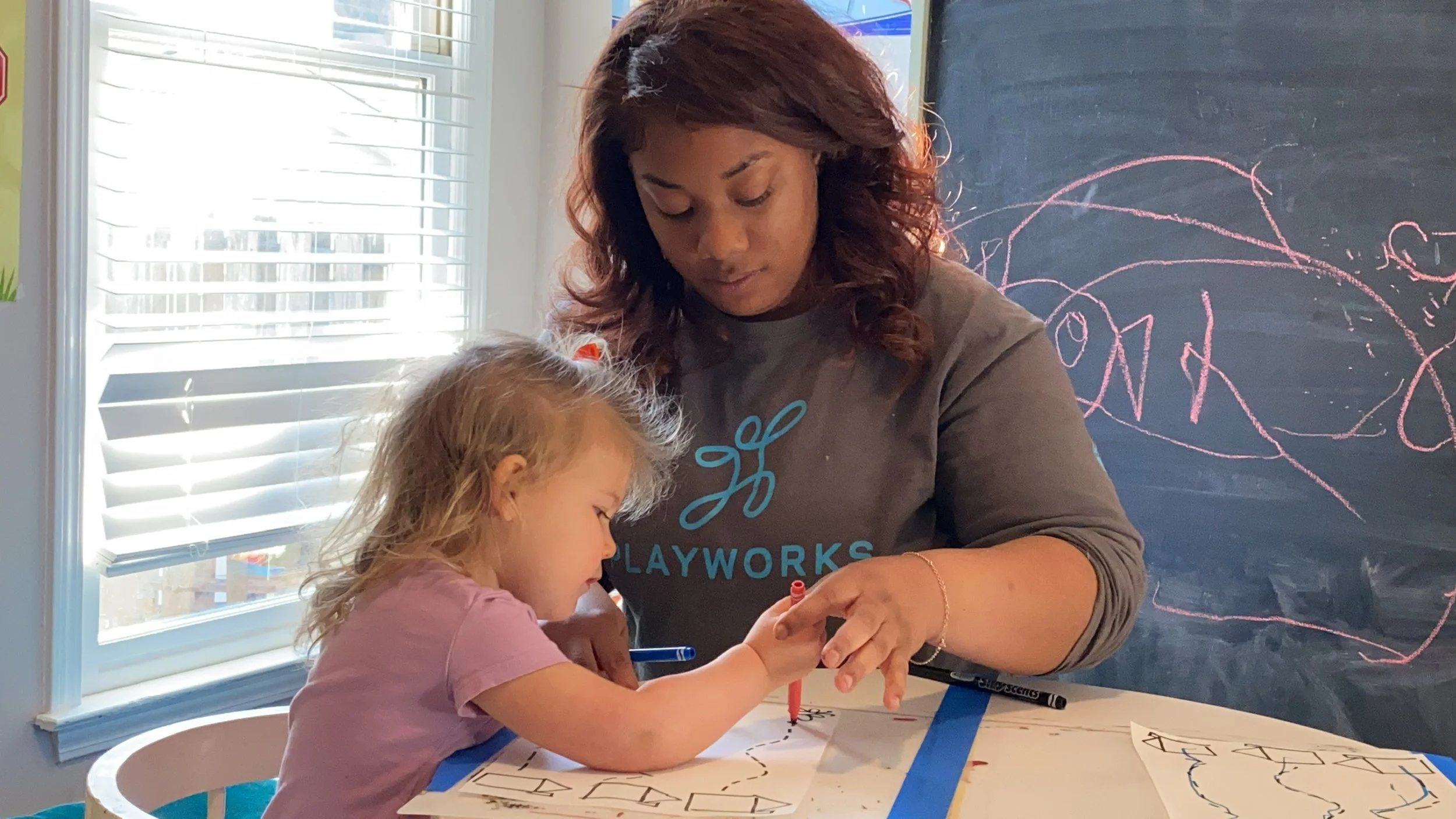 A young girl and a woman with curly hair are working together at a table. The girl is drawing on paper with markers, and the woman is helping or guiding her. There are drawings and scribbles on the paper and on a black chalkboard behind them. The scene is well-lit by natural light coming from a window with blinds.