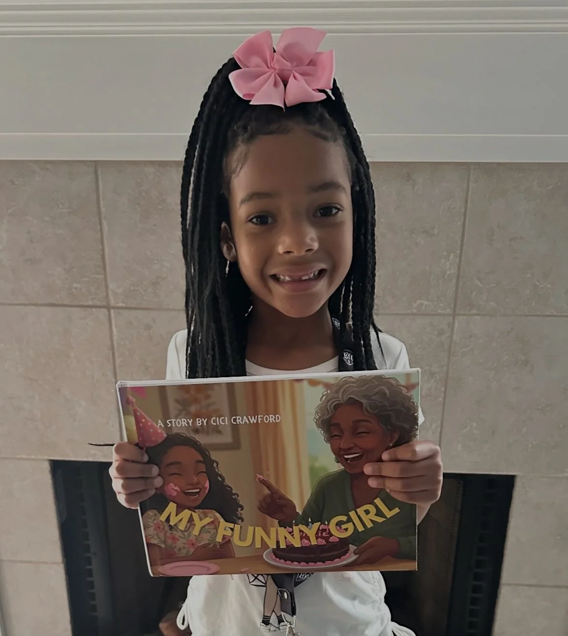 A young girl with dark braided hair and a large pink bow, smiling and holding a children's book titled "My Funny Girl" in front of a beige-tiled wall.
