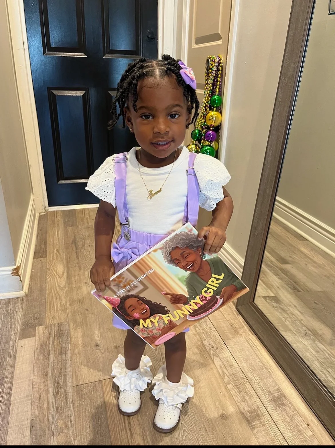 A young girl standing indoors, holding a comic book titled 'My Funny Girl'. She has curly hair decorated with a purple bow, is wearing a white top with lace sleeves, purple suspenders, white ruffled socks, and white shoes. Behind her is a black door and a mirror, with beads and ornaments hanging on the wall.