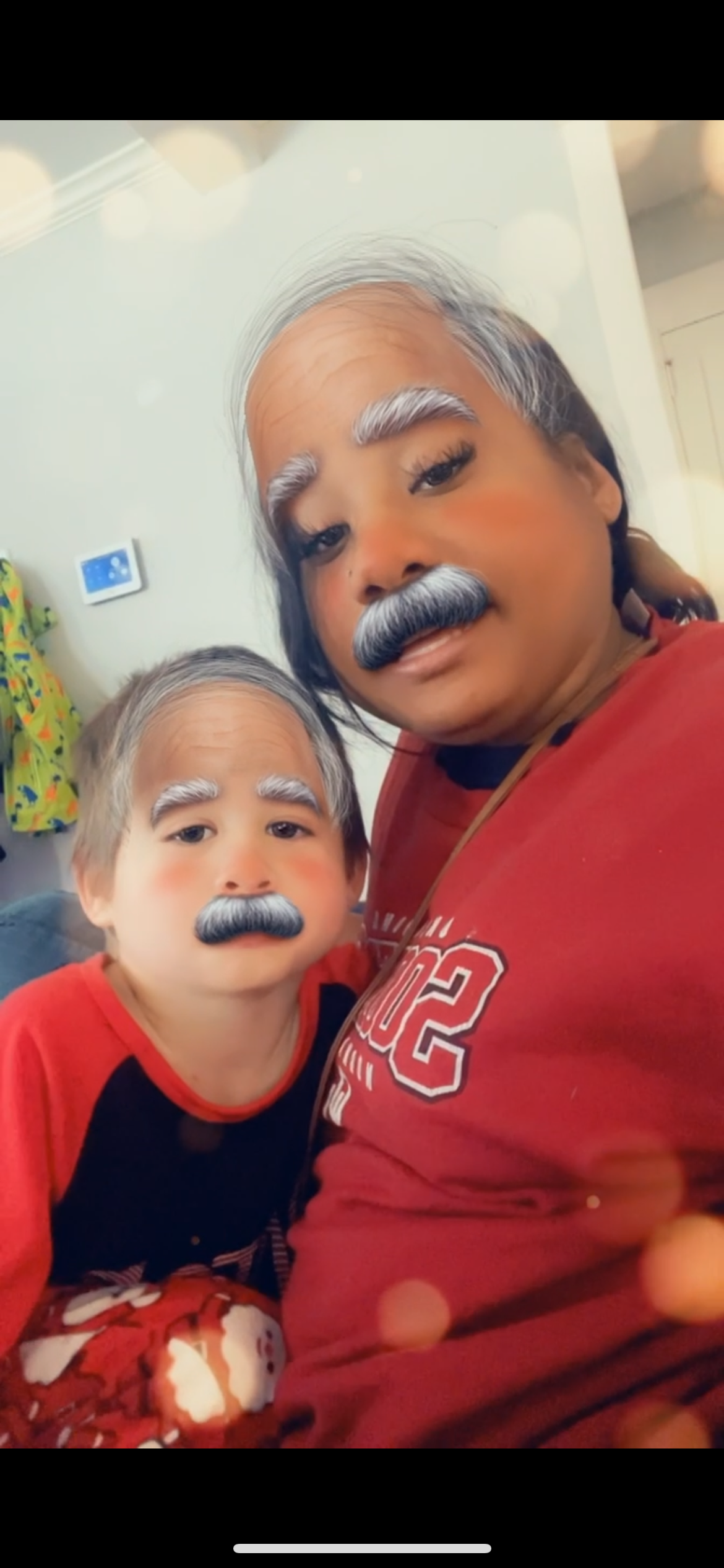 A woman and a young boy taking a selfie together, both with a filter that gives them gray hair, eyebrows, mustaches, and rosy cheeks, resembling elderly figures.