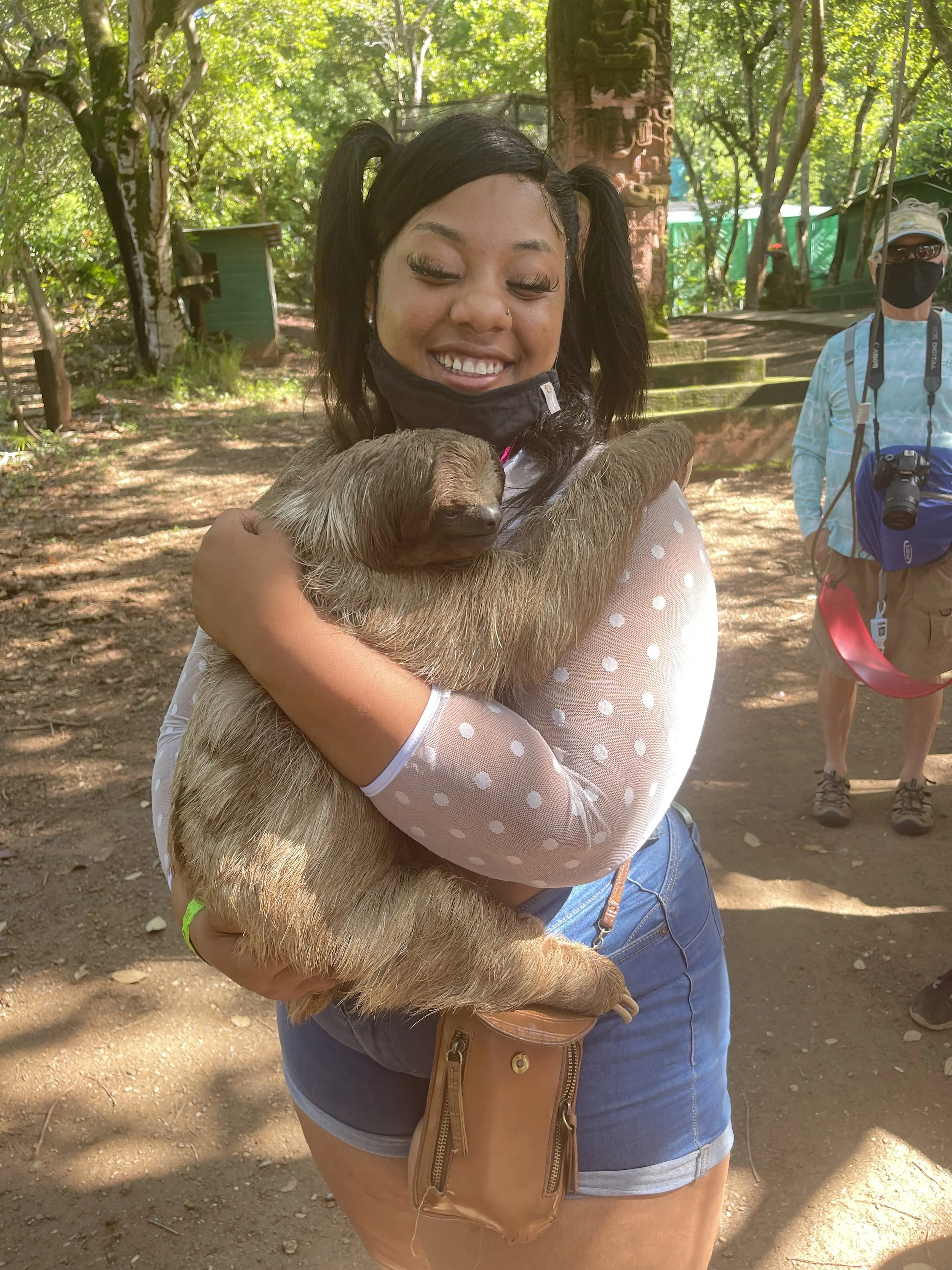 Young woman smiling and hugging a sleeping sloth outdoors in a lush, green forest setting, with a man in the background wearing a face mask and camera gear.