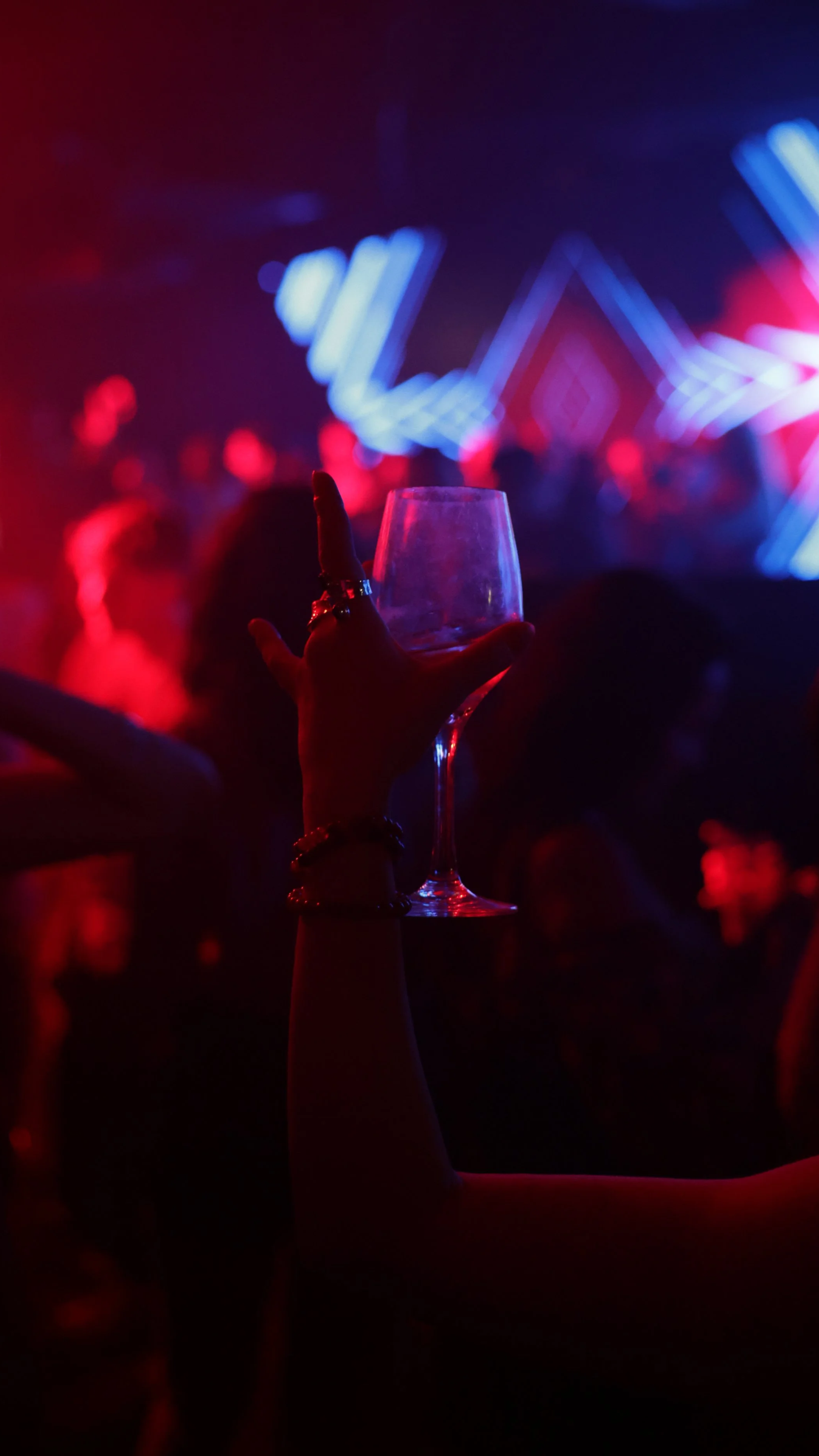 Eine Hand hält ein Weinglas in einem dunklen, bunten Partyraum mit Neonlichtern und tanzenden Menschen im Hintergrund.