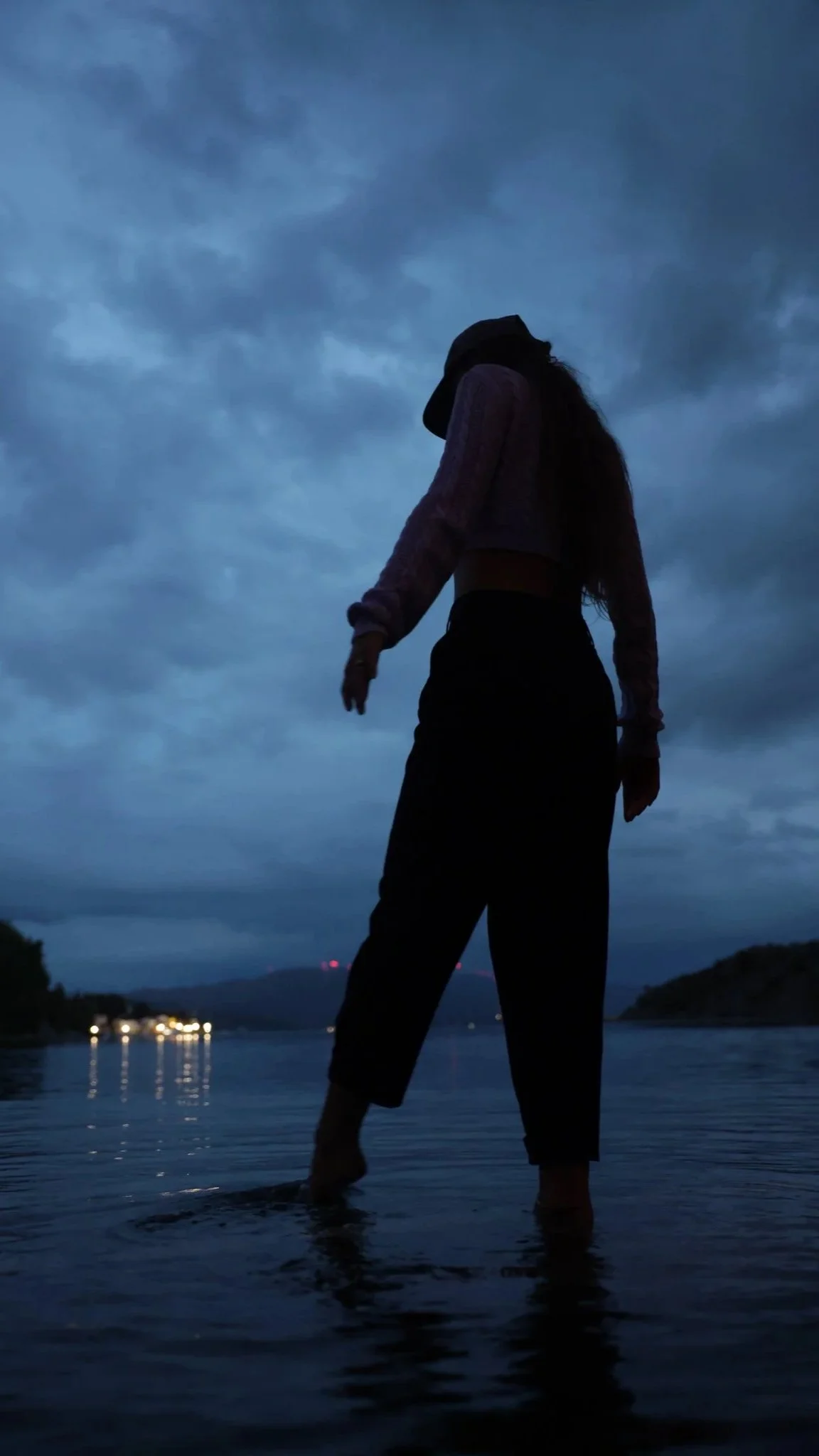 Eine silhouettierte Person steht im Wasser bei Dämmerung, mit dunklem Himmel und Wolken im Hintergrund.