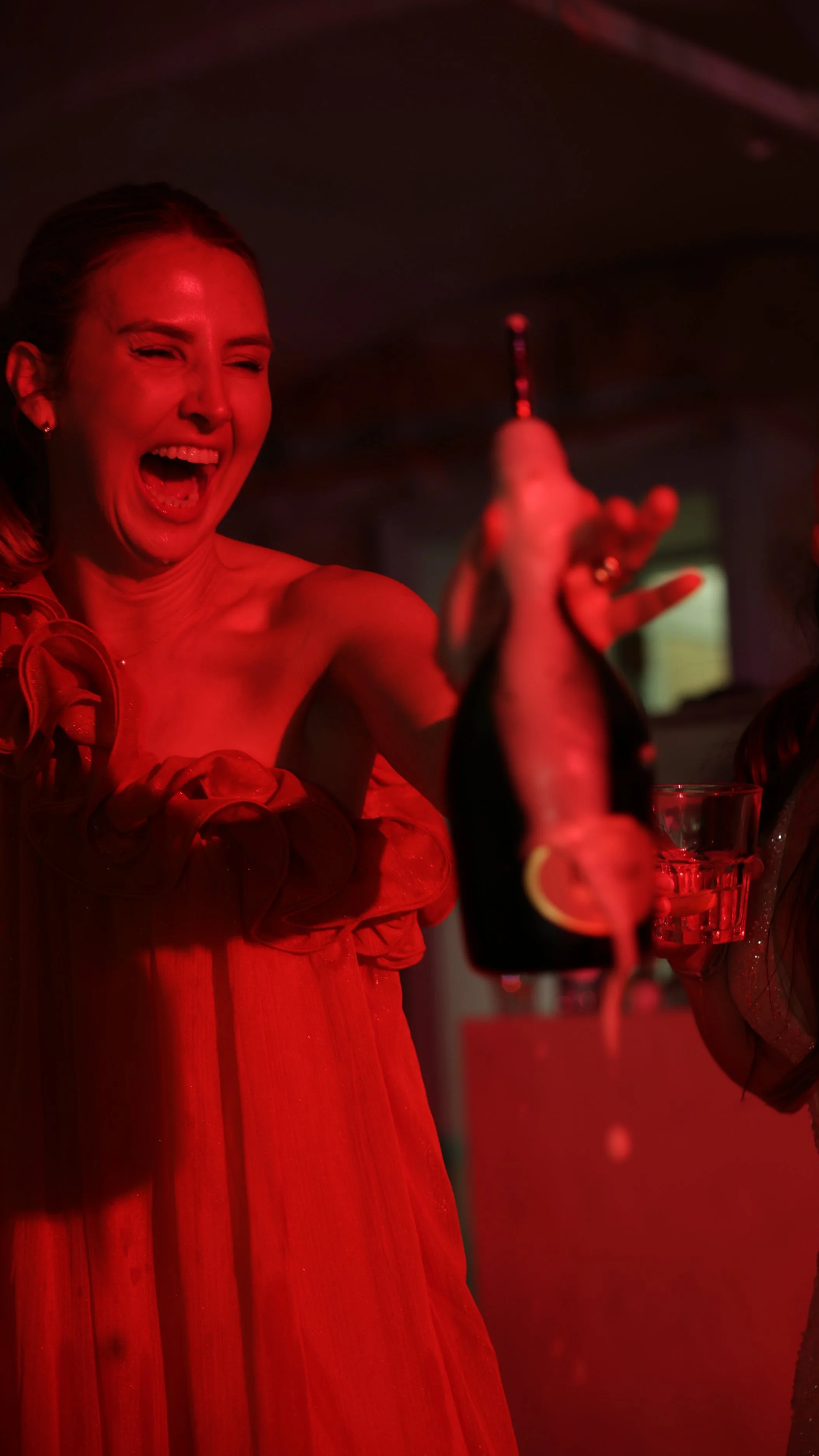 Frau in rotem Abendkleid hält eine Flasche Sekt in einer Bar, lacht fröhlich.