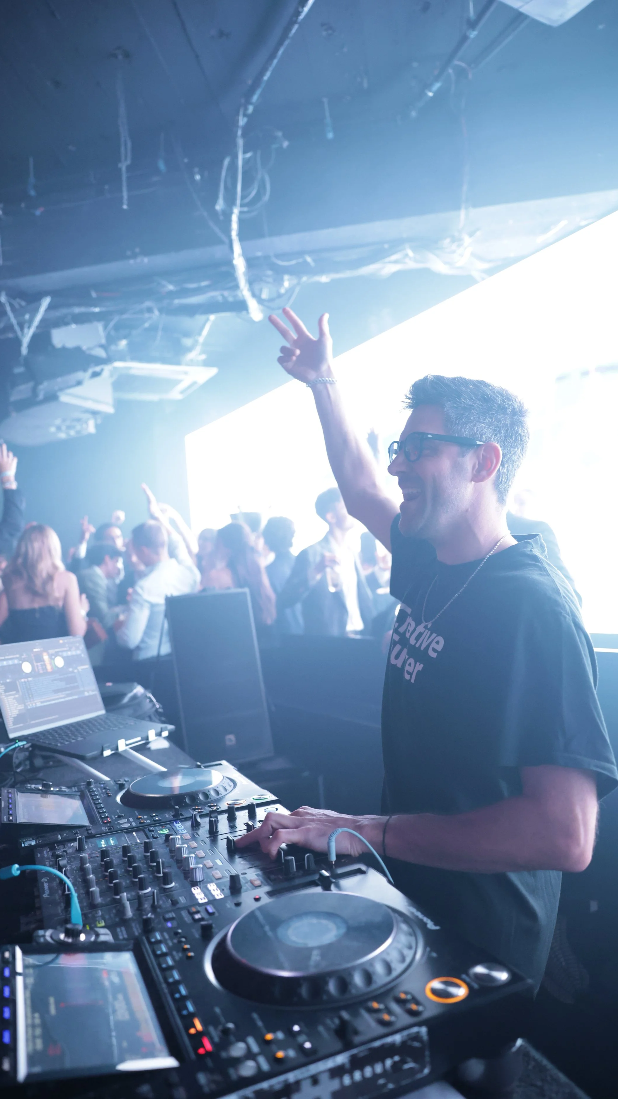 DJ spielt Musik in einer clubartigen Umgebung mit tanzenden Menschen im Hintergrund, die vor einem großen helleuchten Bildschirm stehen.