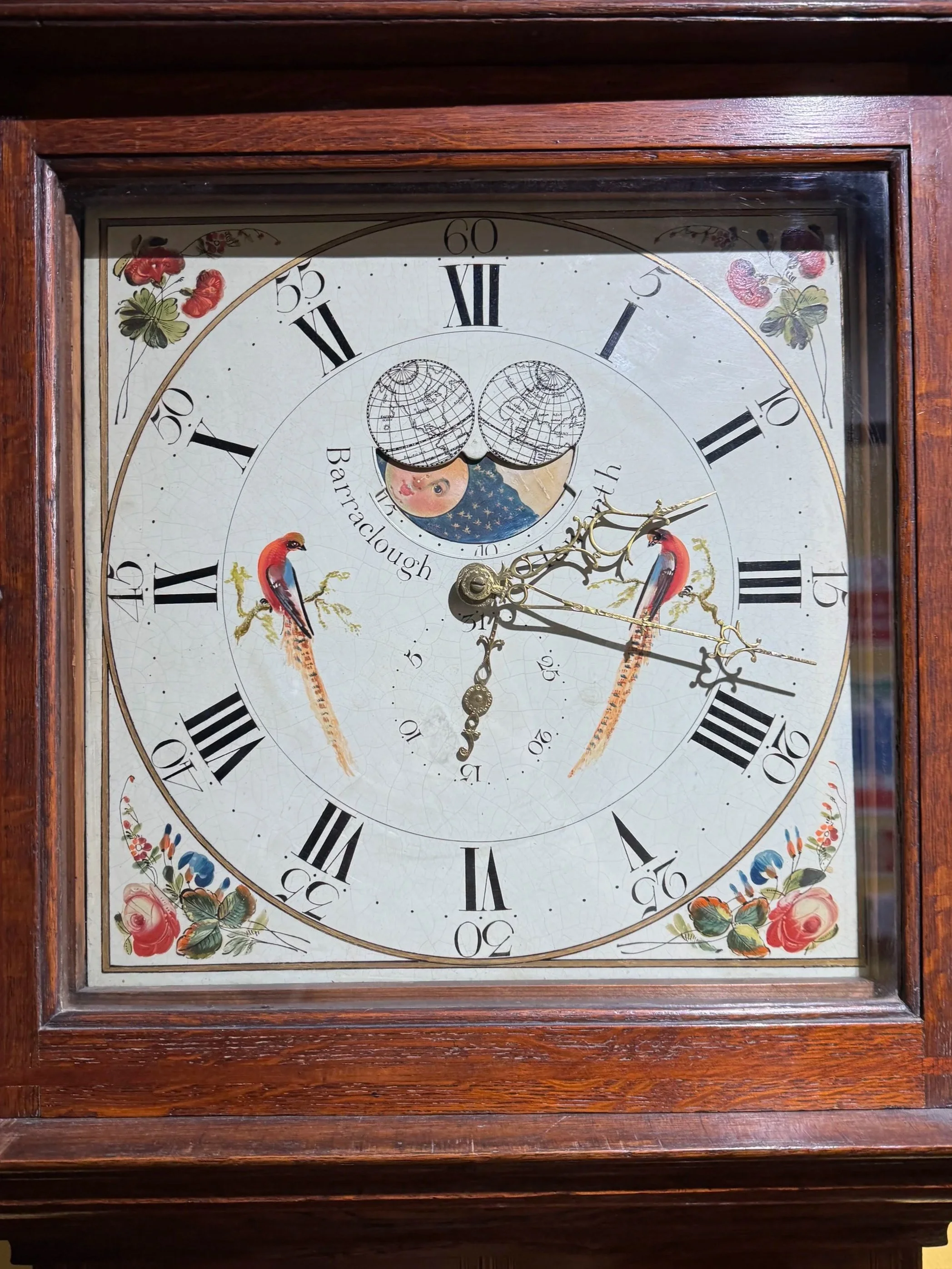 An antique wooden clock with a decorative face showing hours in Roman numerals and featuring images of two pinkish-red cranes, a moon with a child's face, and floral accents.