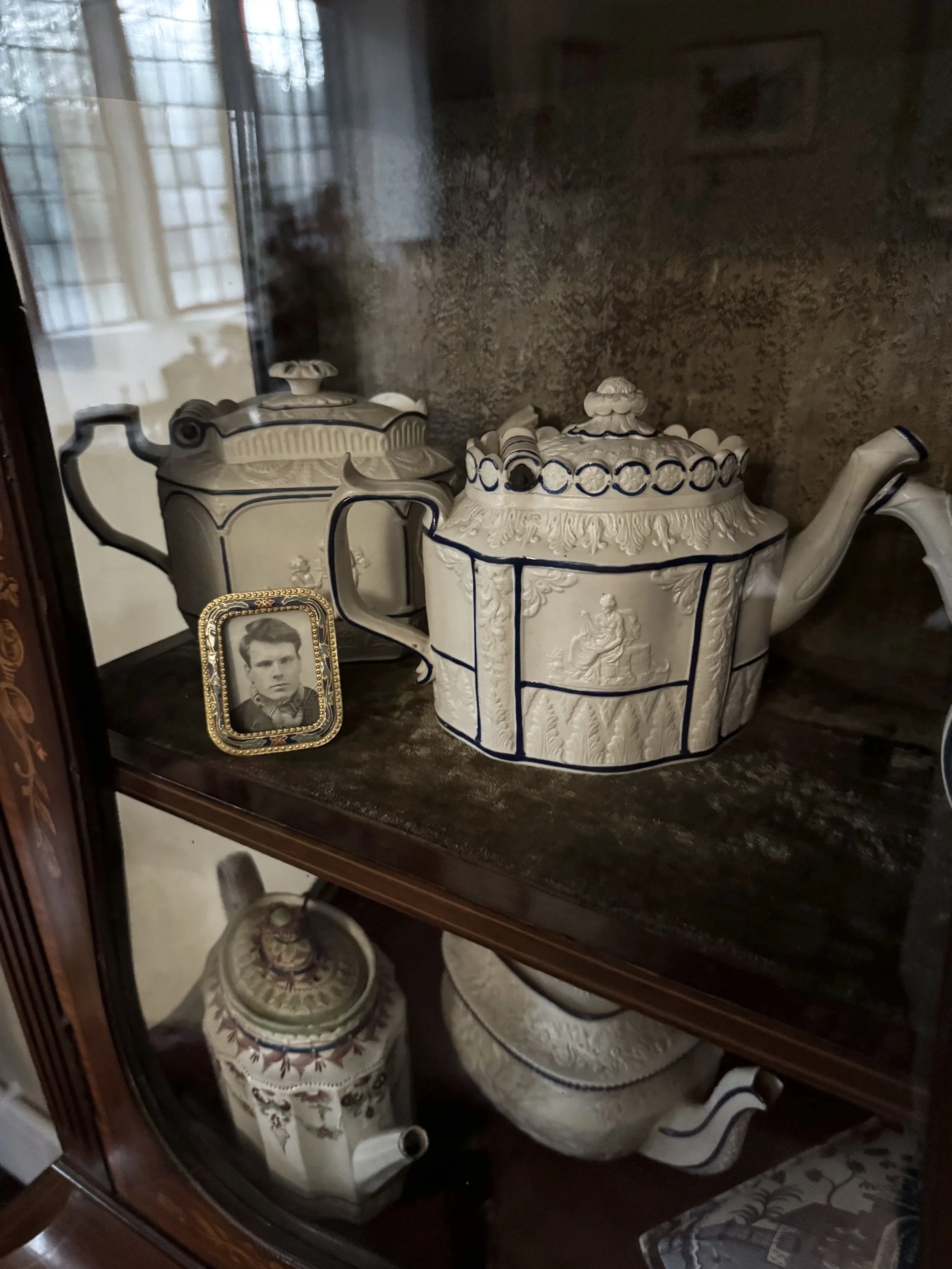 Antique porcelain tea set with a teapot and a sugar bowl, a small black-and-white photograph, and other decorative items inside a glass display cabinet.
