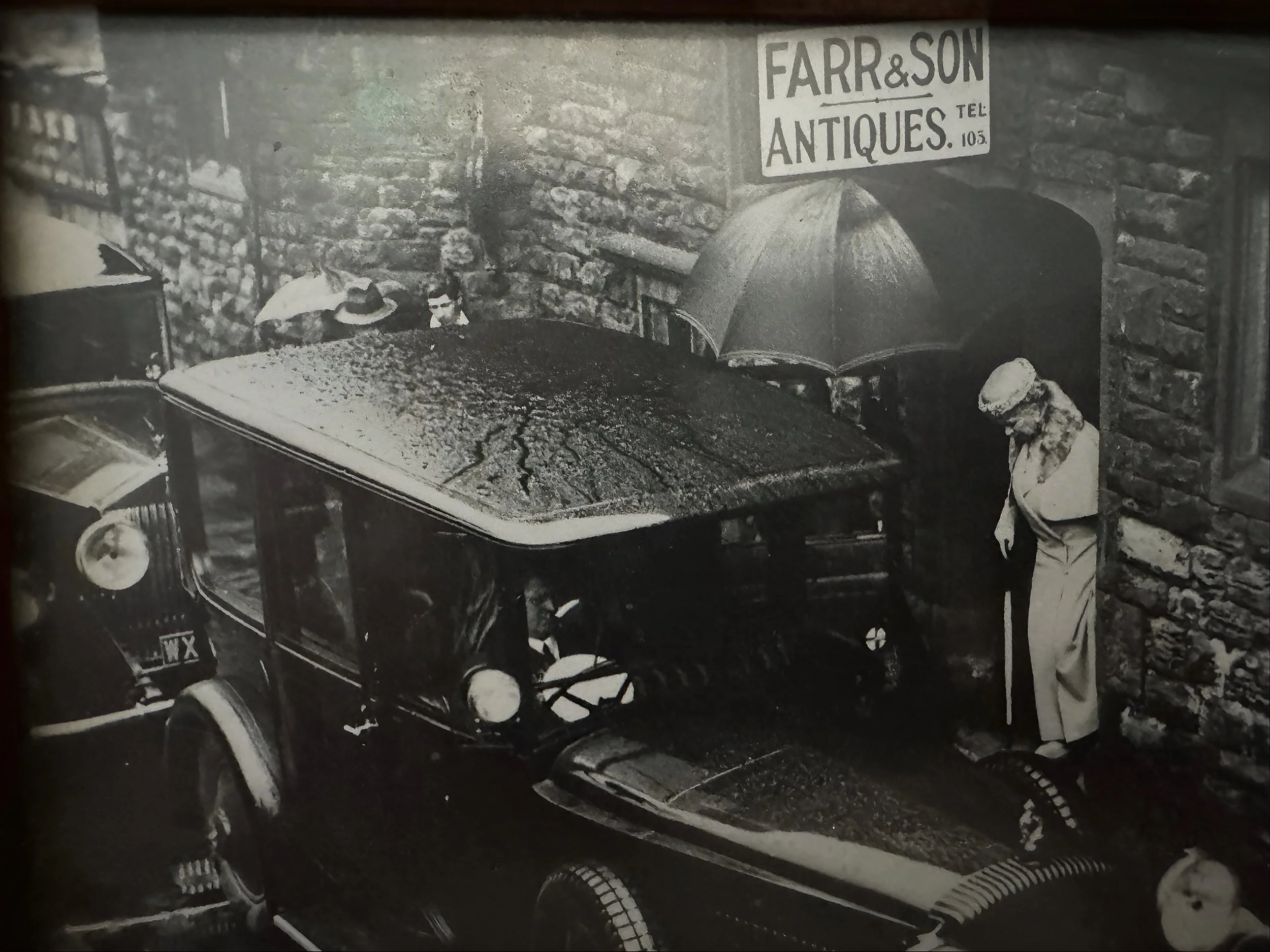 Black and white photo showing a vintage car parked on a cobblestone street, with a woman wearing a hat and dress standing next to its hood. A woman holding an umbrella is also visible near the car, and a sign for 'FARR & SON ANTIQUES' is mounted on a brick wall above an arched doorway.