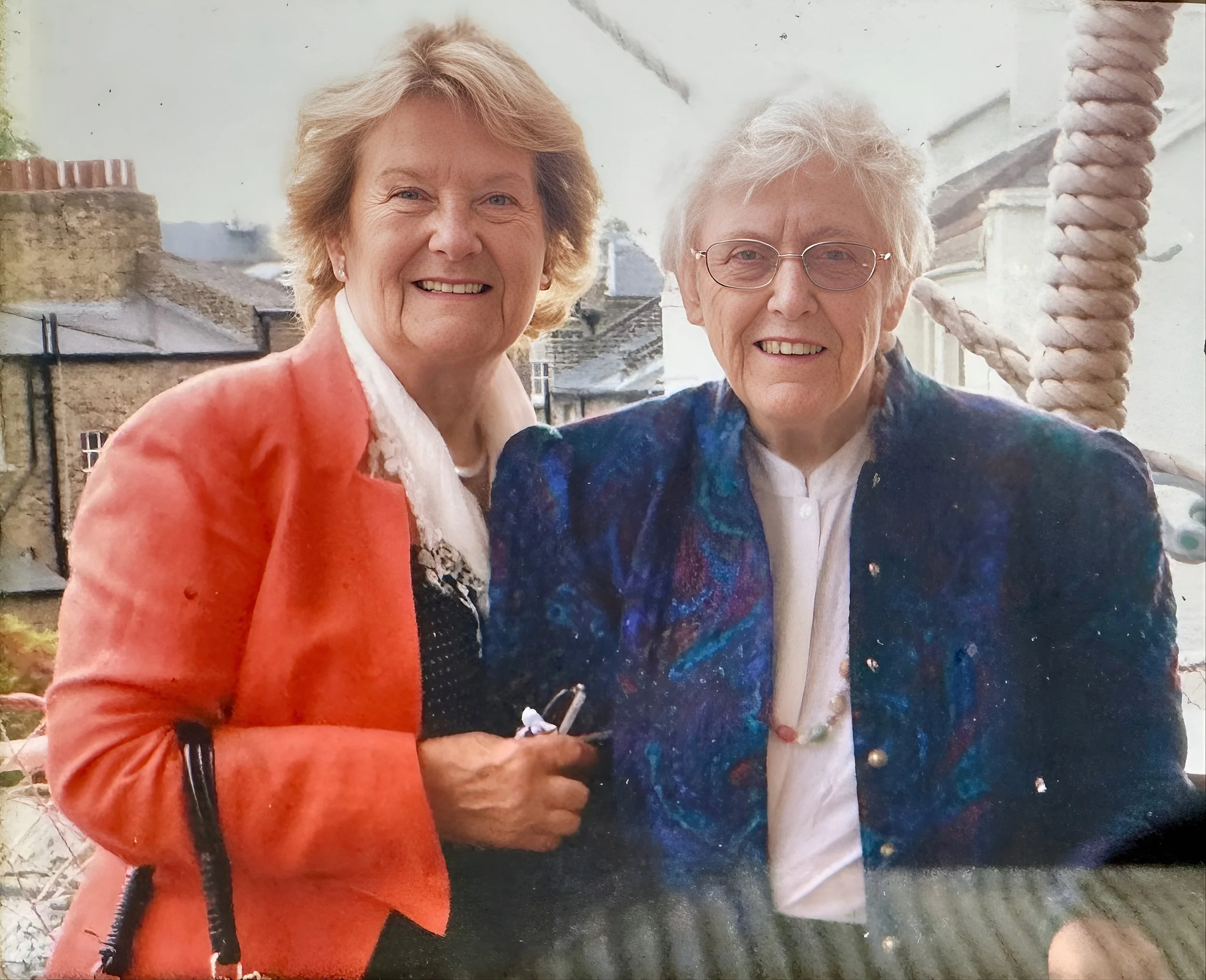 Two elderly women smiling outdoors, one with short blonde hair wearing a coral jacket and the other with short white hair and glasses wearing a blue patterned jacket, standing close together with positive expressions.