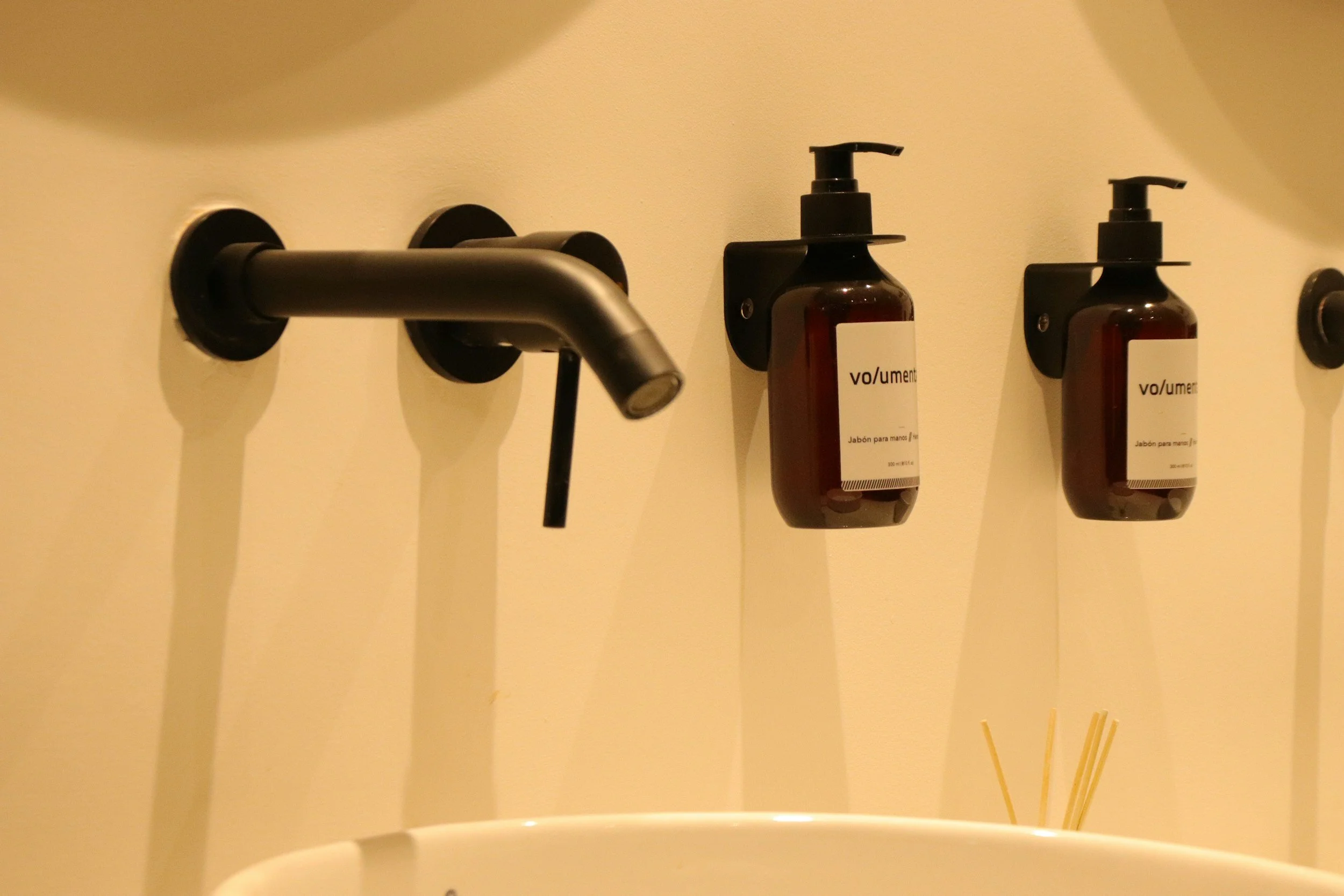 A wall-mounted black faucet with a downward spout, two amber soap dispensers labeled 'vo/umem', and a white ceramic sink or basin below, with a bundle of reed sticks to the side.