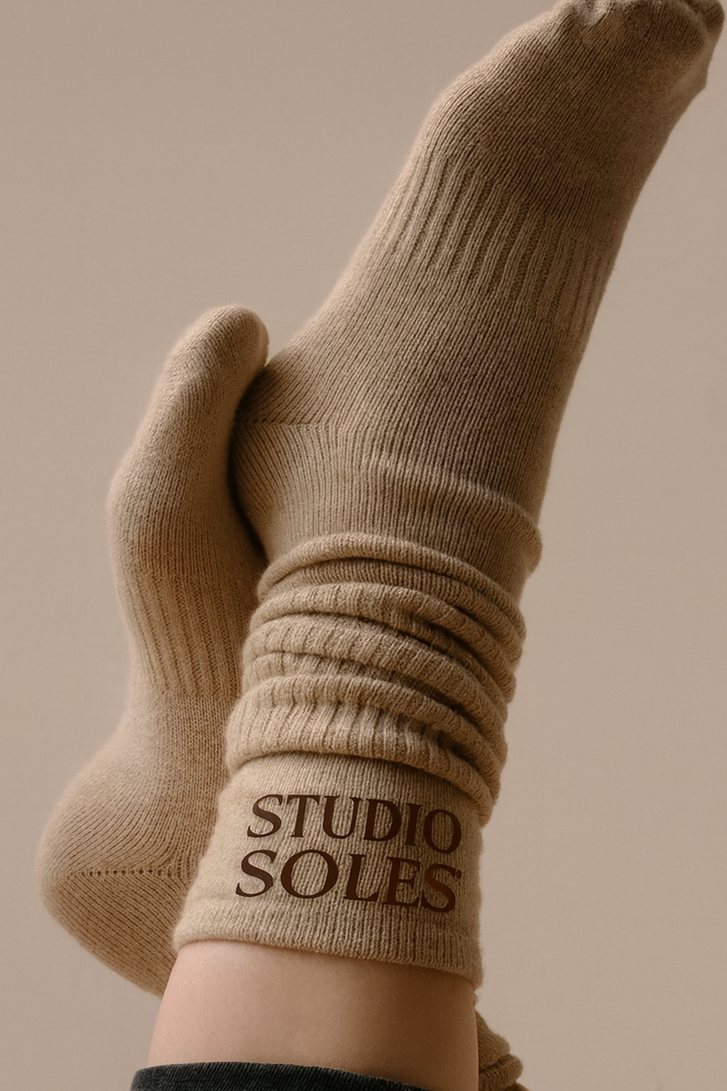 A person's foot in a tan sock with the words 'STUDIO SOLES' written on the cuff, held up against a neutral background.