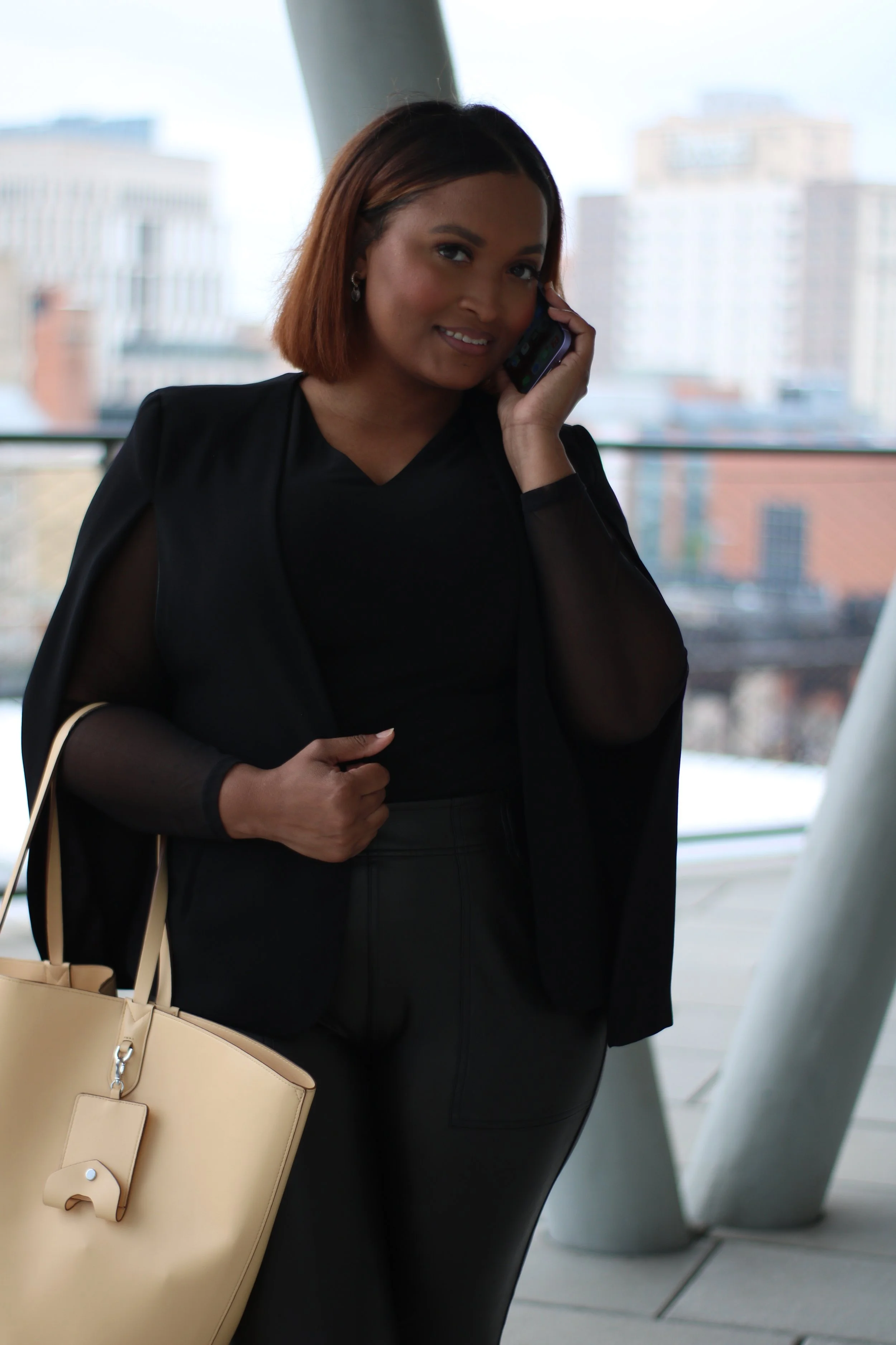 A woman with shoulder-length brown hair talking on a cellphone while standing in an indoor architectural space with large windows and city buildings in the background.