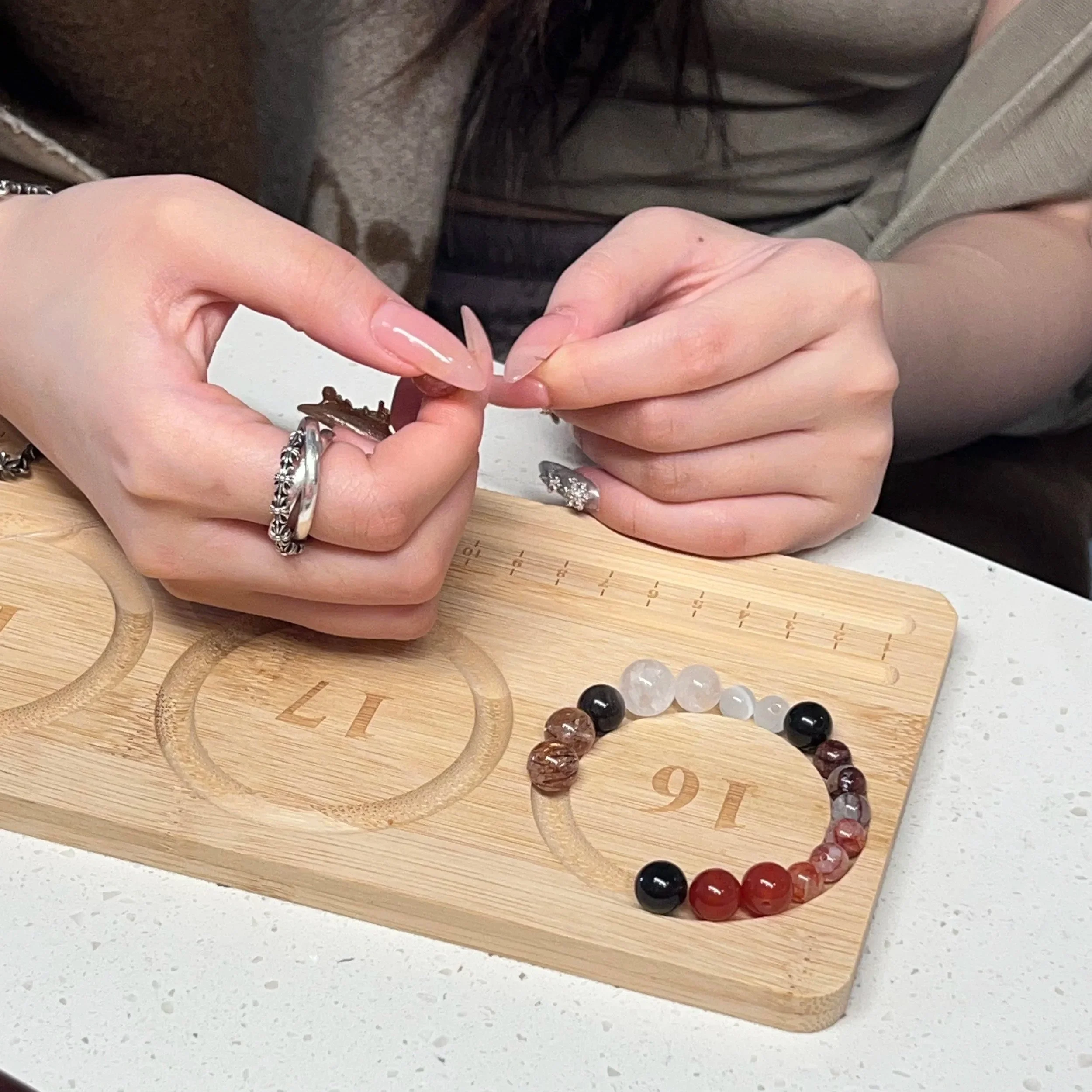 A person wearing rings on their fingers is stringing beads onto a bracelet. The beads are circular and multicolored, placed on a wooden bead board with labeled circles.