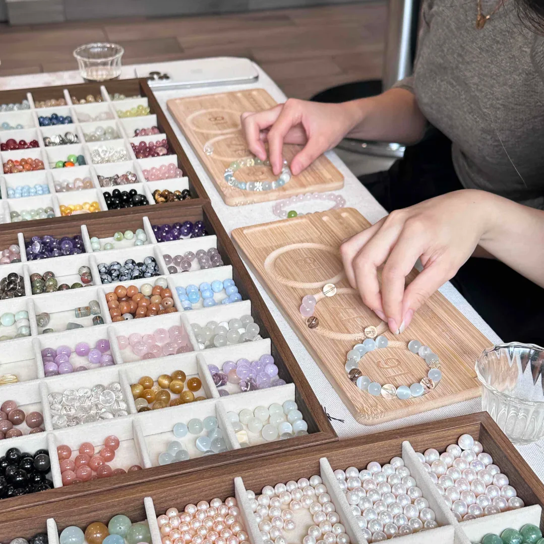 Person arranging pearl and gemstone bracelets on wooden display boards surrounded by boxes of various colored beads.