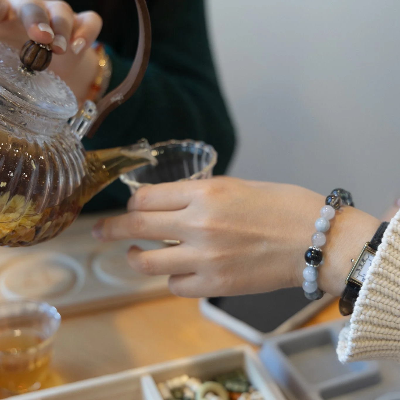 Person pouring tea from a glass teapot into a glass cup, with a hand wearing a watch and bracelet visible, on a wooden table with some food and a smartphone in the background.