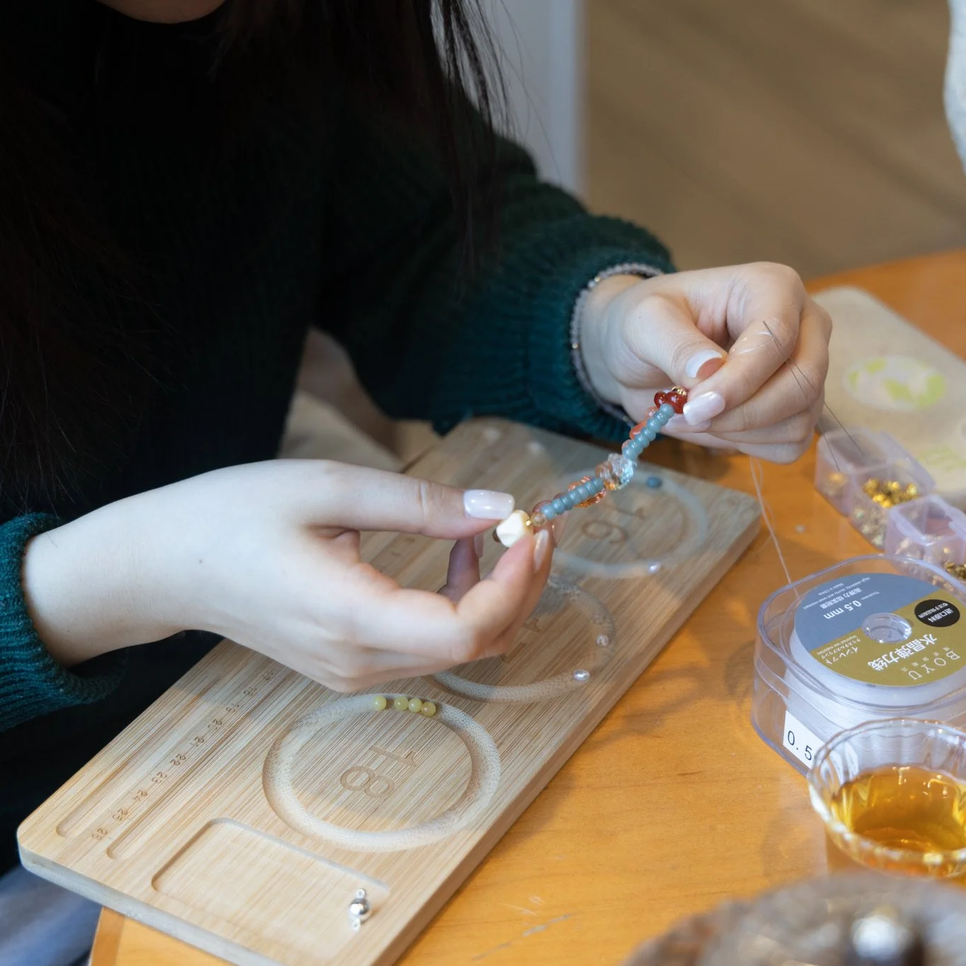 A person making a beaded bracelet using colorful beads, sitting at a wooden table with jewelry-making supplies.