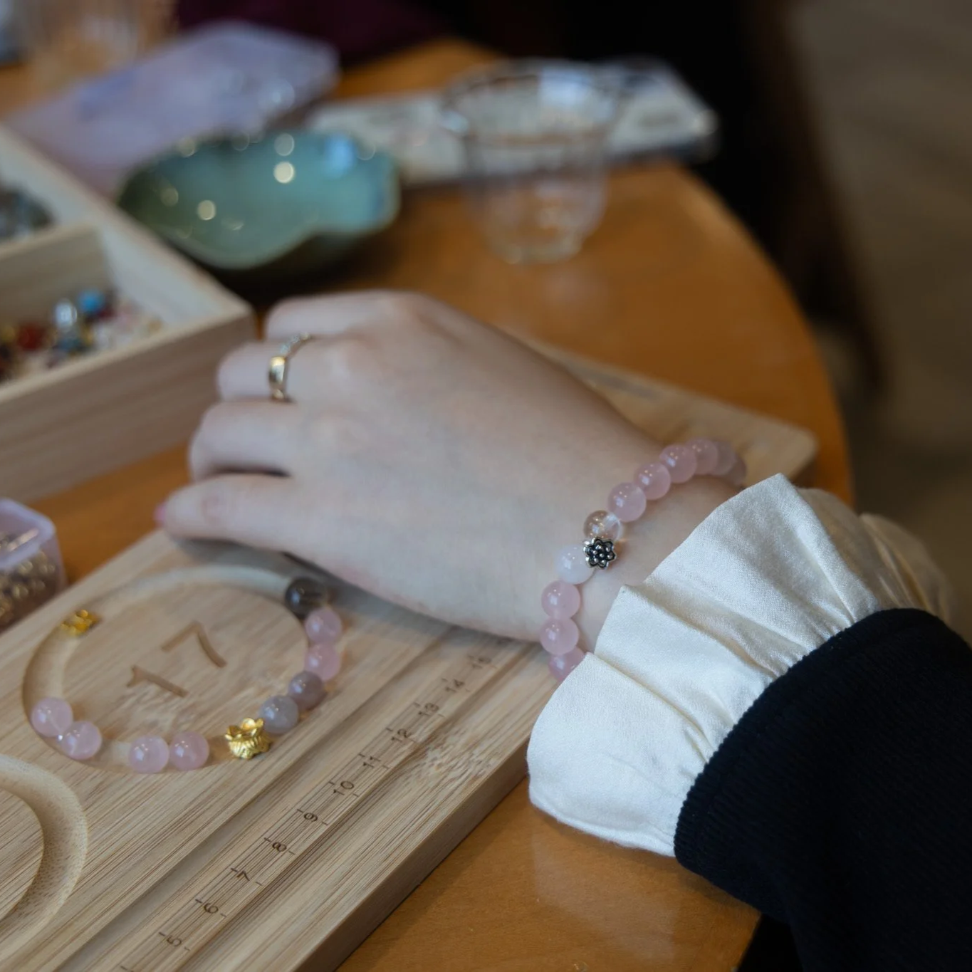 A person's hand resting on a wooden display board with jewelry, including a pink beaded bracelet and matching necklace, surrounded by jewelry boxes on a wooden table.